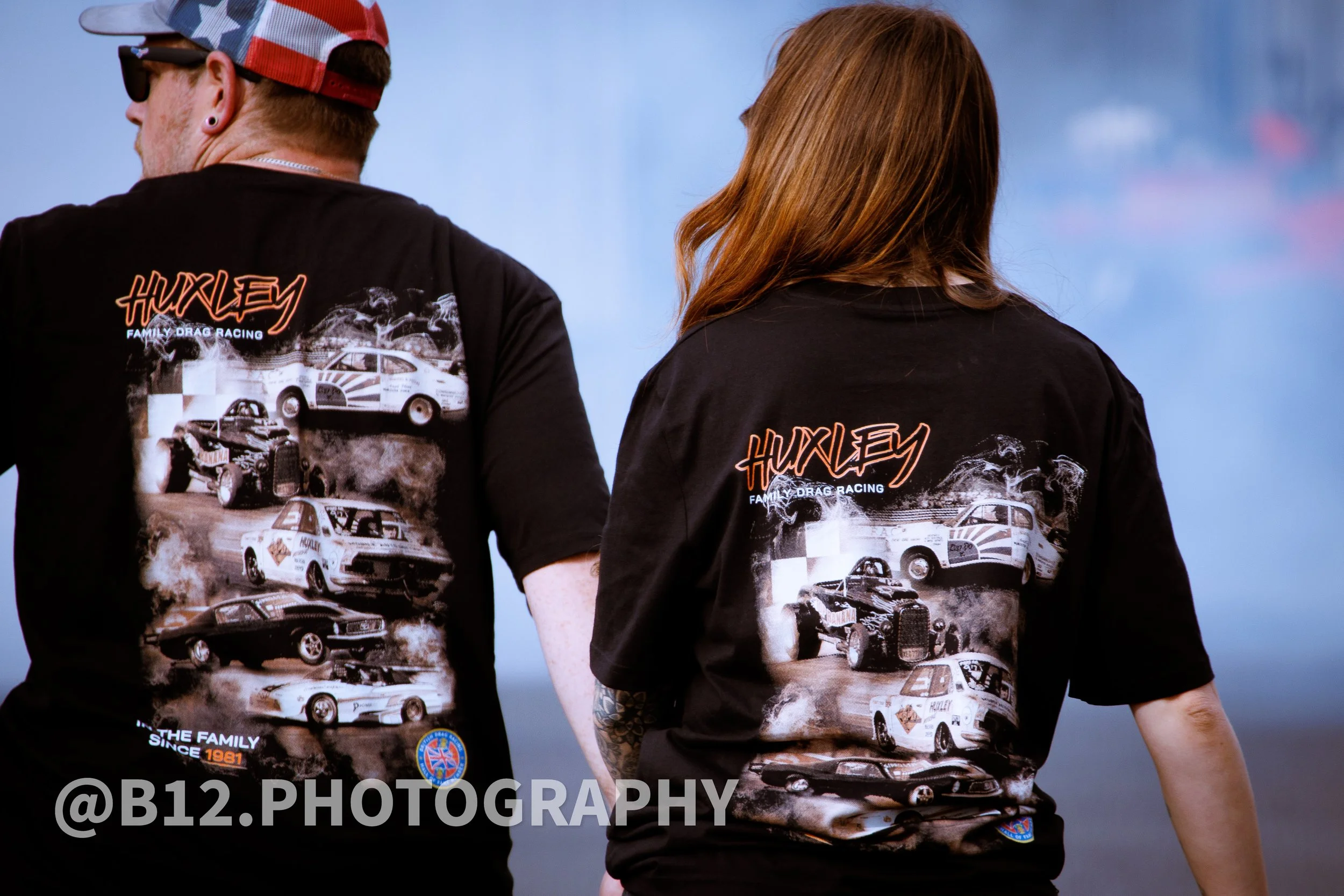 Two people wearing black T-shirts with car and racing graphics, standing outdoors against a blurred background.