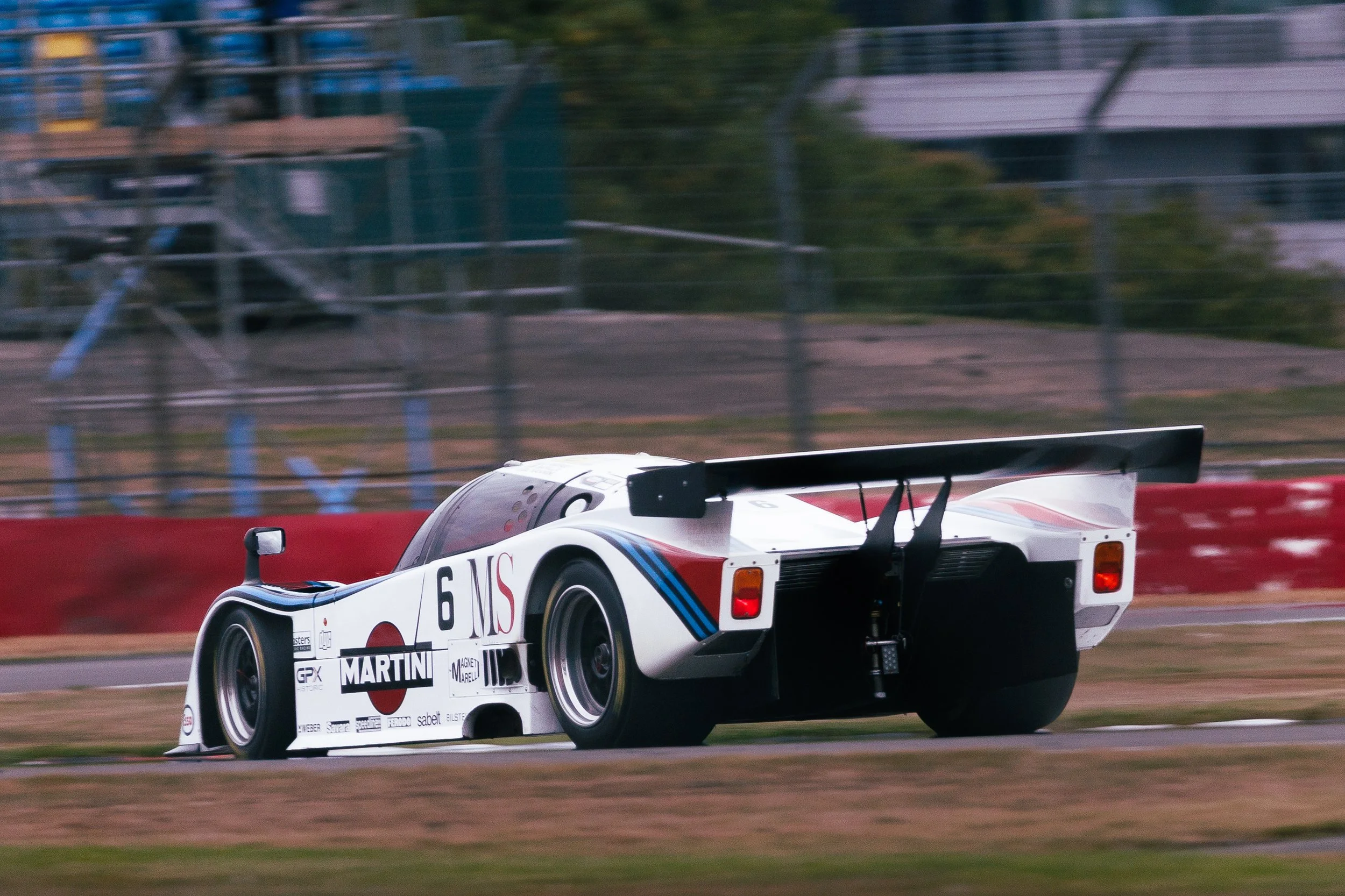 A white vintage race car with sponsorship logos and the number 6 on its side is speeding on a racetrack with a blurred background of fencing and trees.