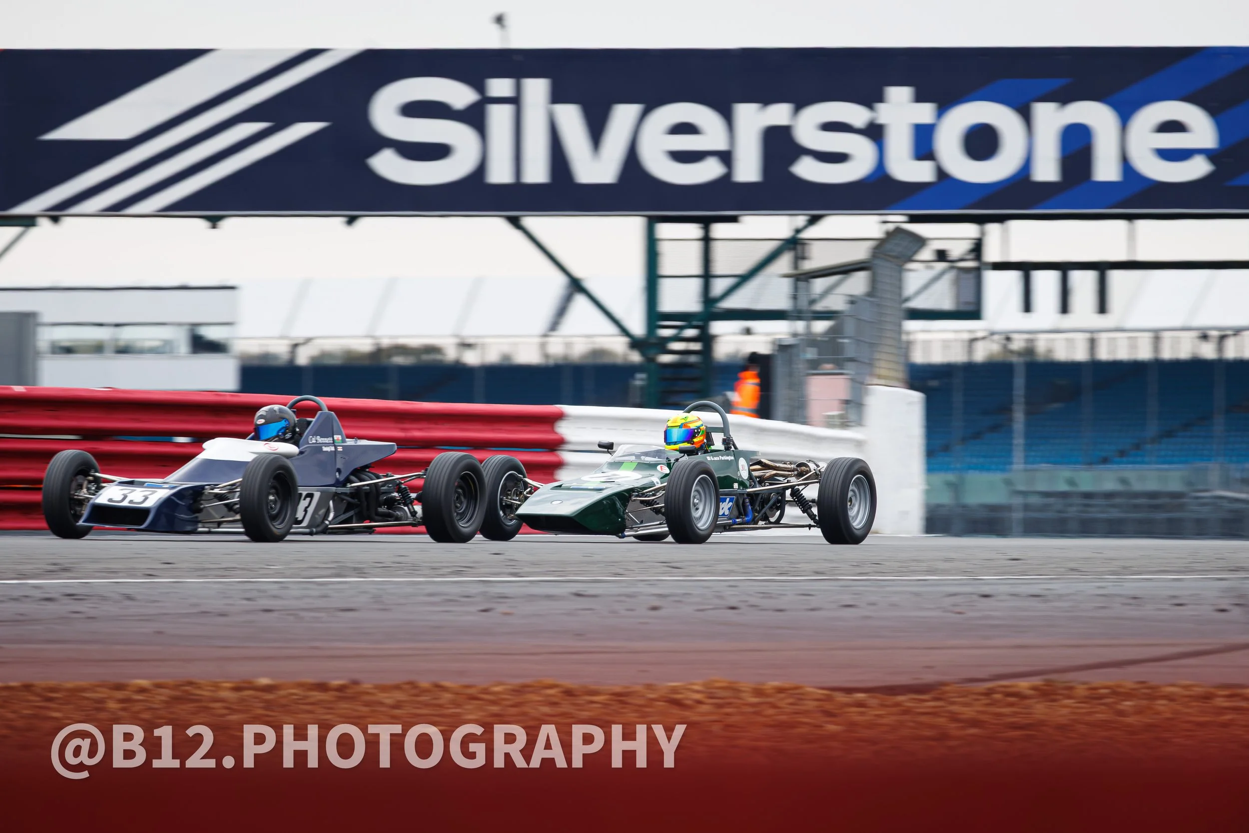 Two vintage race cars on a track, with one car bumping into the other. The background displays a large sign that says 'Silverstone'.