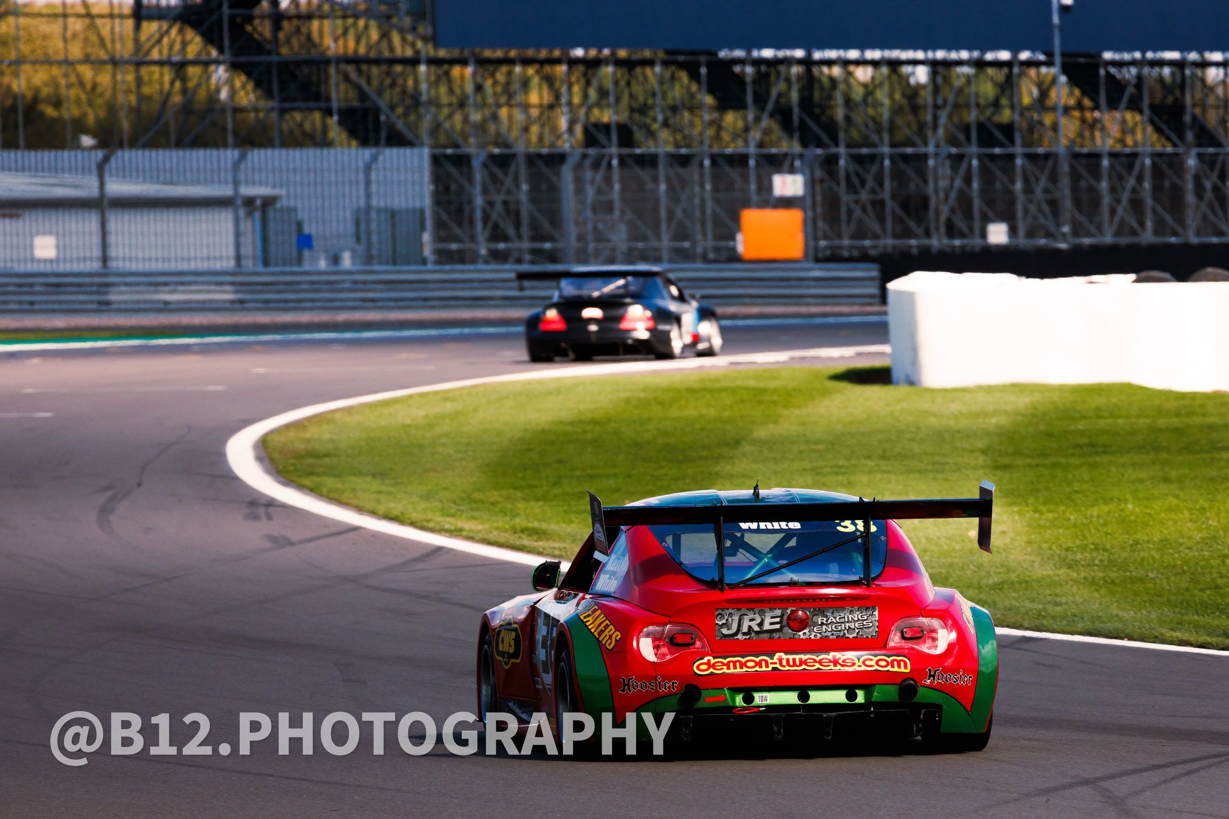 A red and green race car on a racetrack with another black car in the distance and structural barriers in the background.