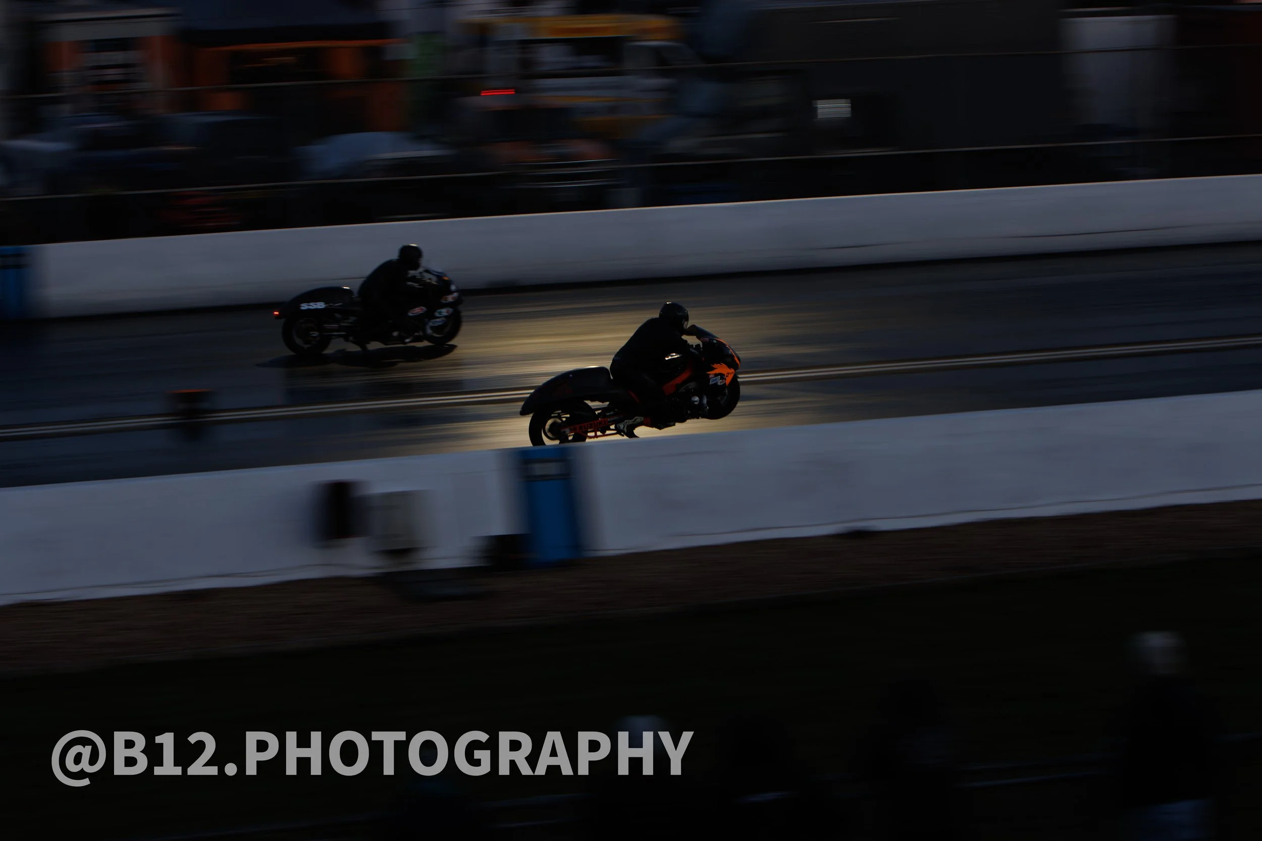 Two motorcycles racing on a wet track during dusk or night with illuminated headlights, with a blurred background of spectators and structures.