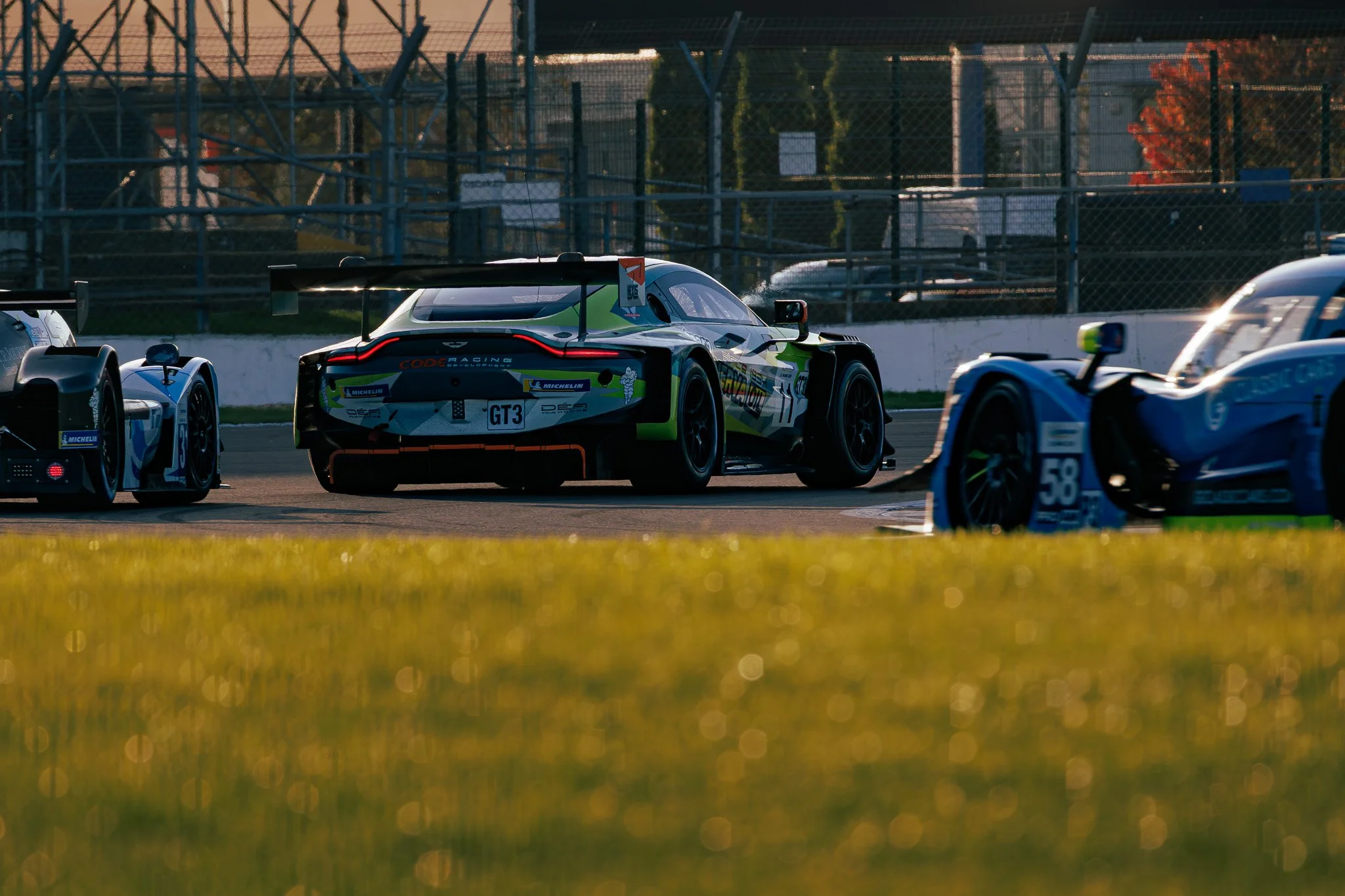 Race cars on a track, with one car in the foreground and others in the background during sunset.