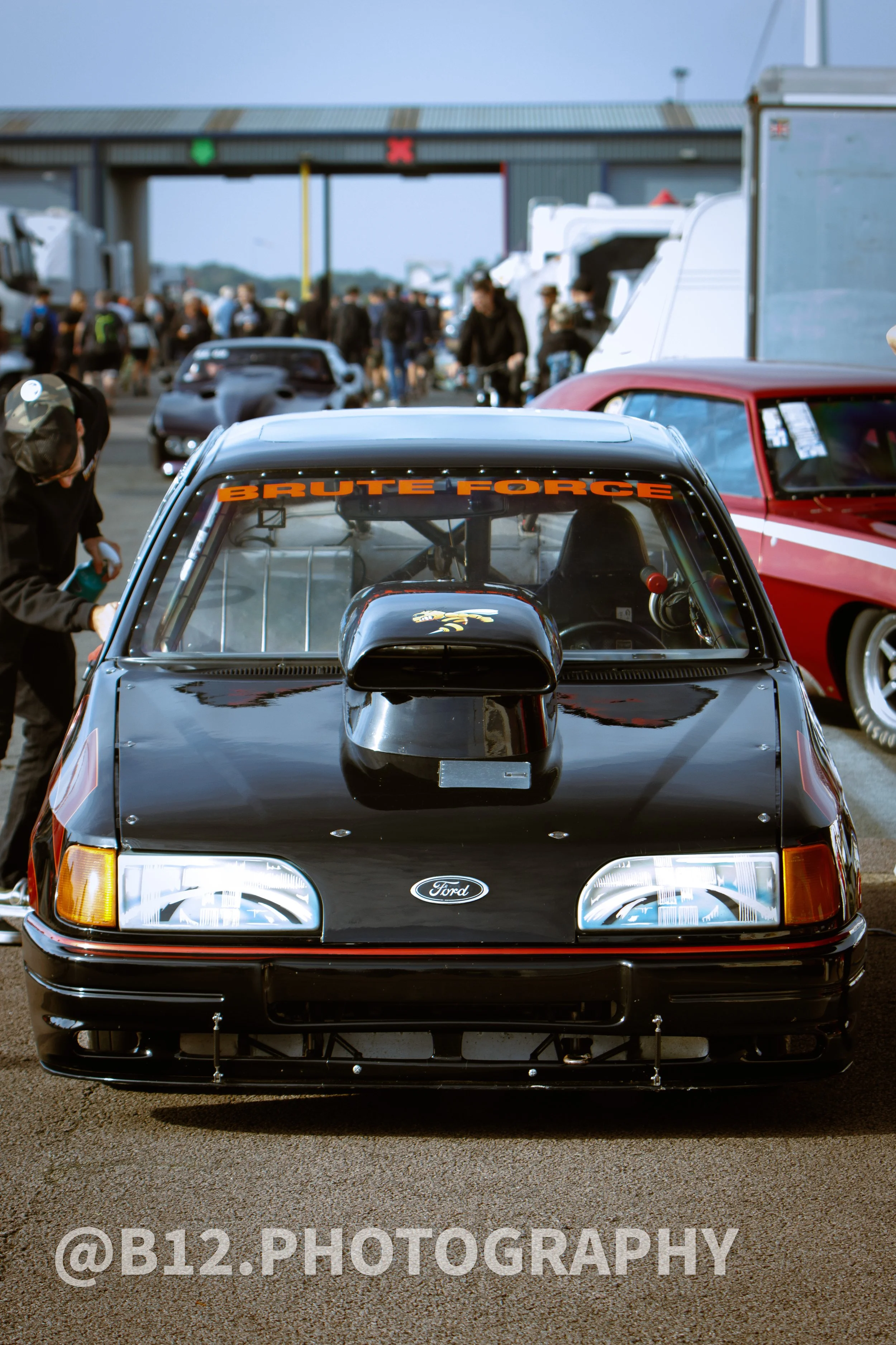 Front view of a black Ford race car with a prominent supercharger on the hood, parked among other cars at a racing event.