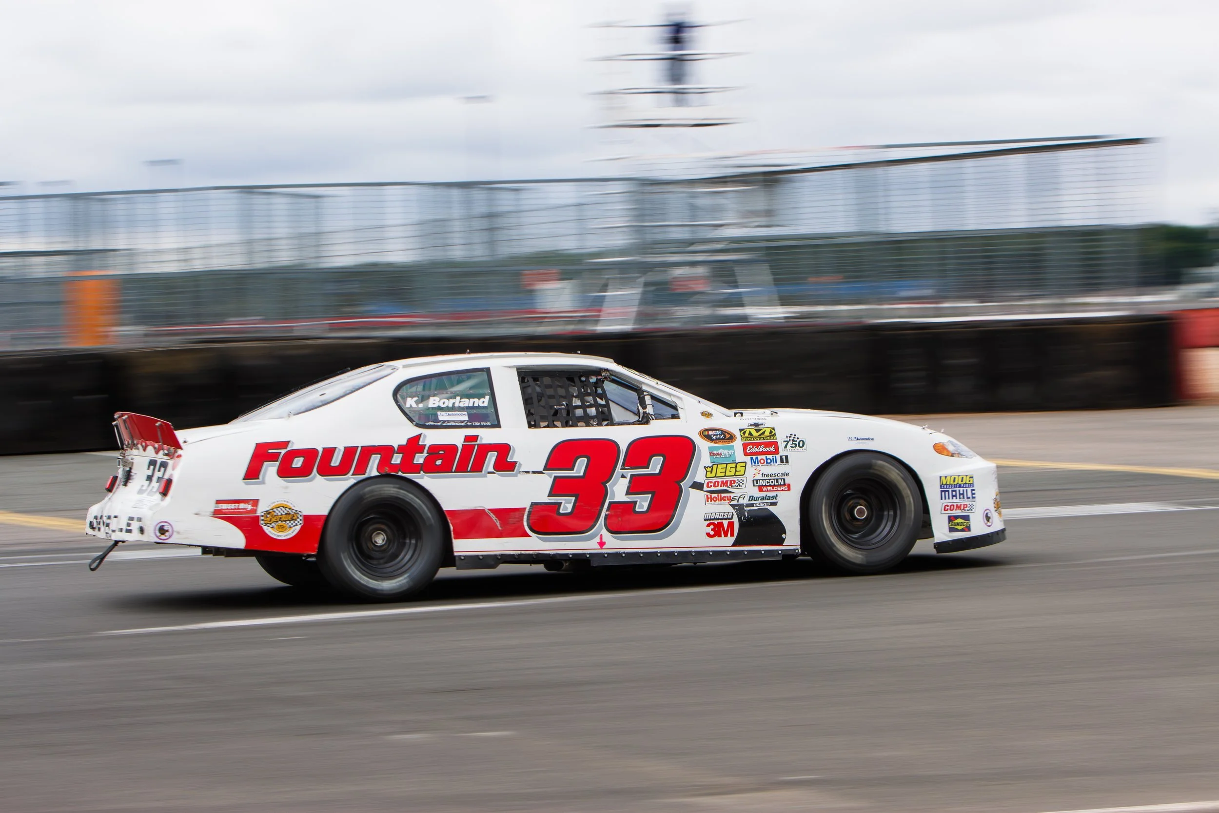 A white race car with red accents, the number 33, and sponsors like Fountain, JEGS, and 3M driving on a racetrack. The driver is K. Borland. The background includes a barrier and blurred spectators.
