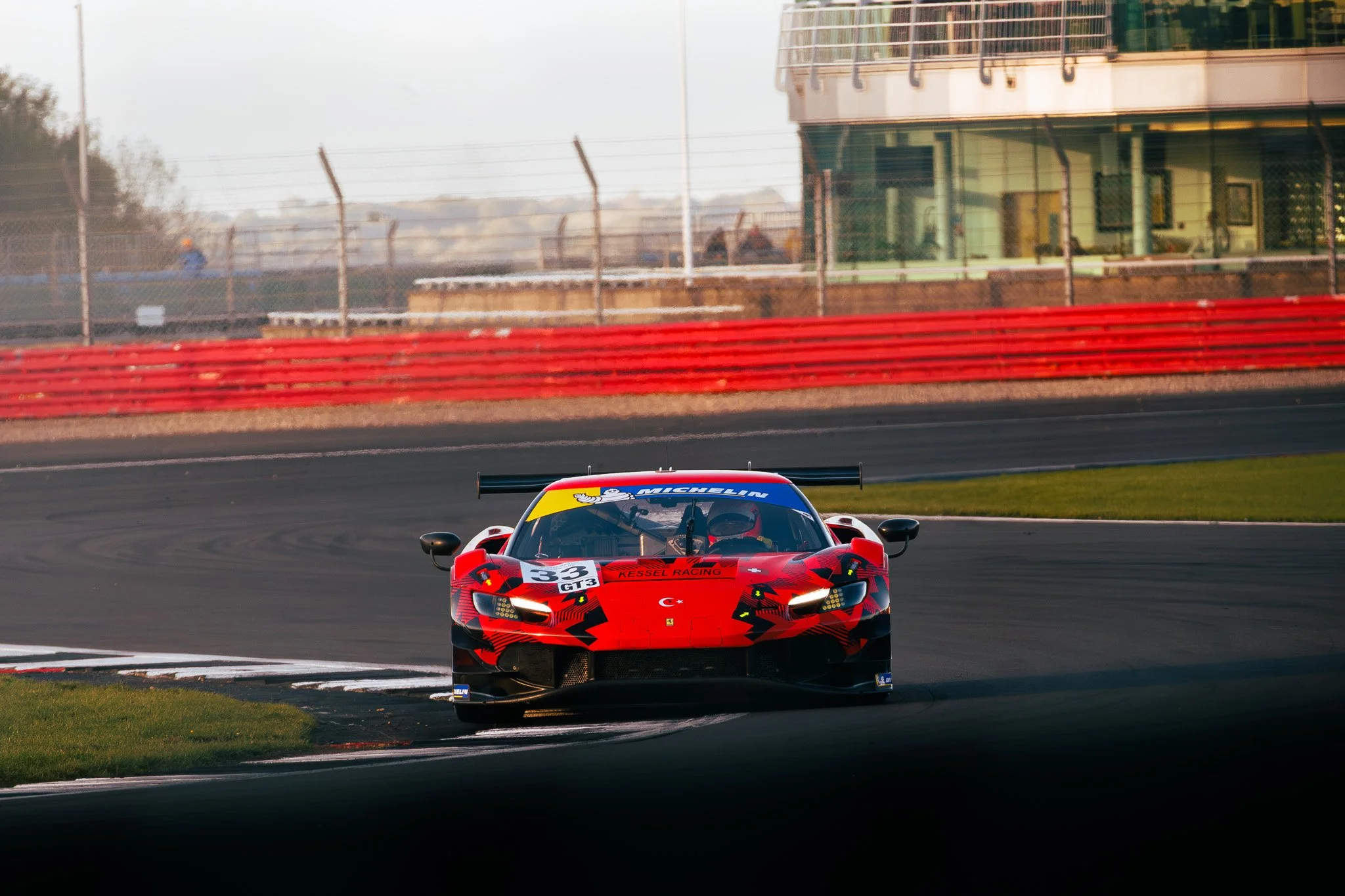 A red race car navigating a turn on a race track, with a building in the background and safety barriers along the side of the track.