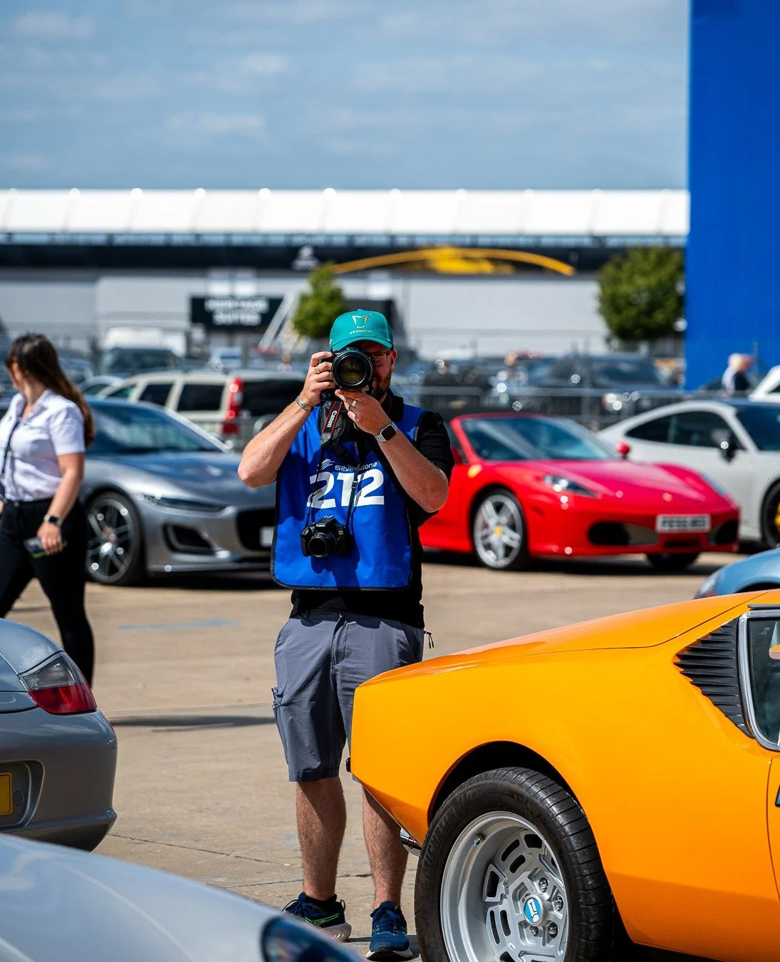 Photographer capturing images at a car event with various sports cars, including an orange one in the foreground, other cars in the background, and a woman walking past.