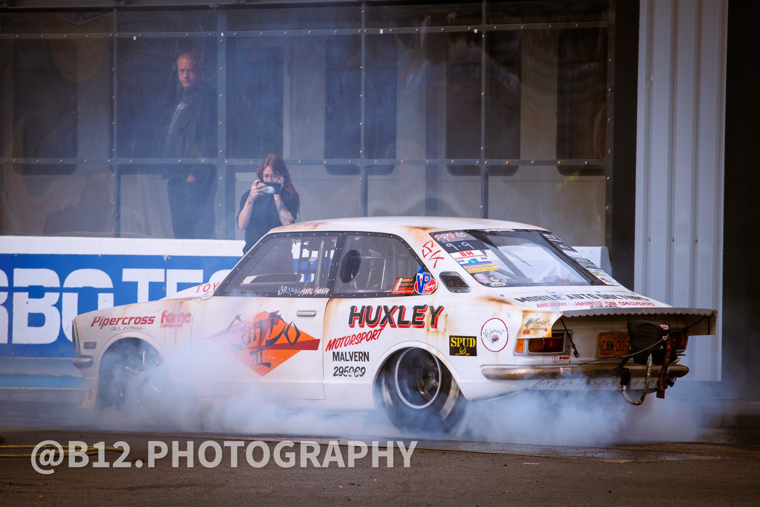 A vintage race car with A/W decals and sponsor logos, smoking tires, in front of a glass enclosure, with two people observing and taking photos.