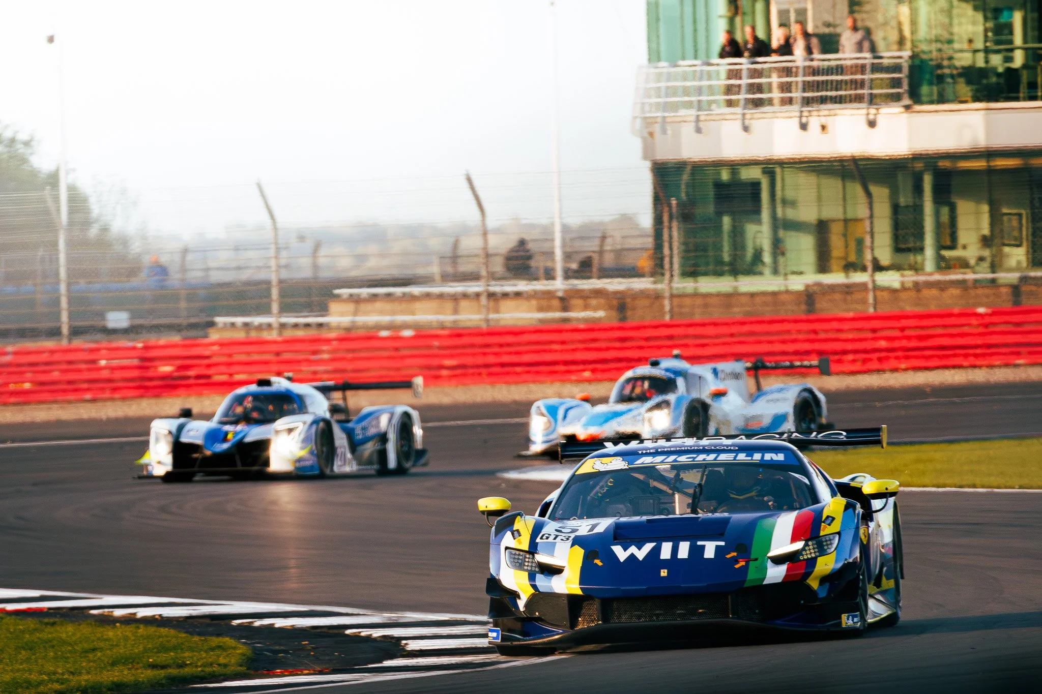 Three race cars on a race track, with the car in the front labeled 'WITT,' followed by two other cars in a racing event.
