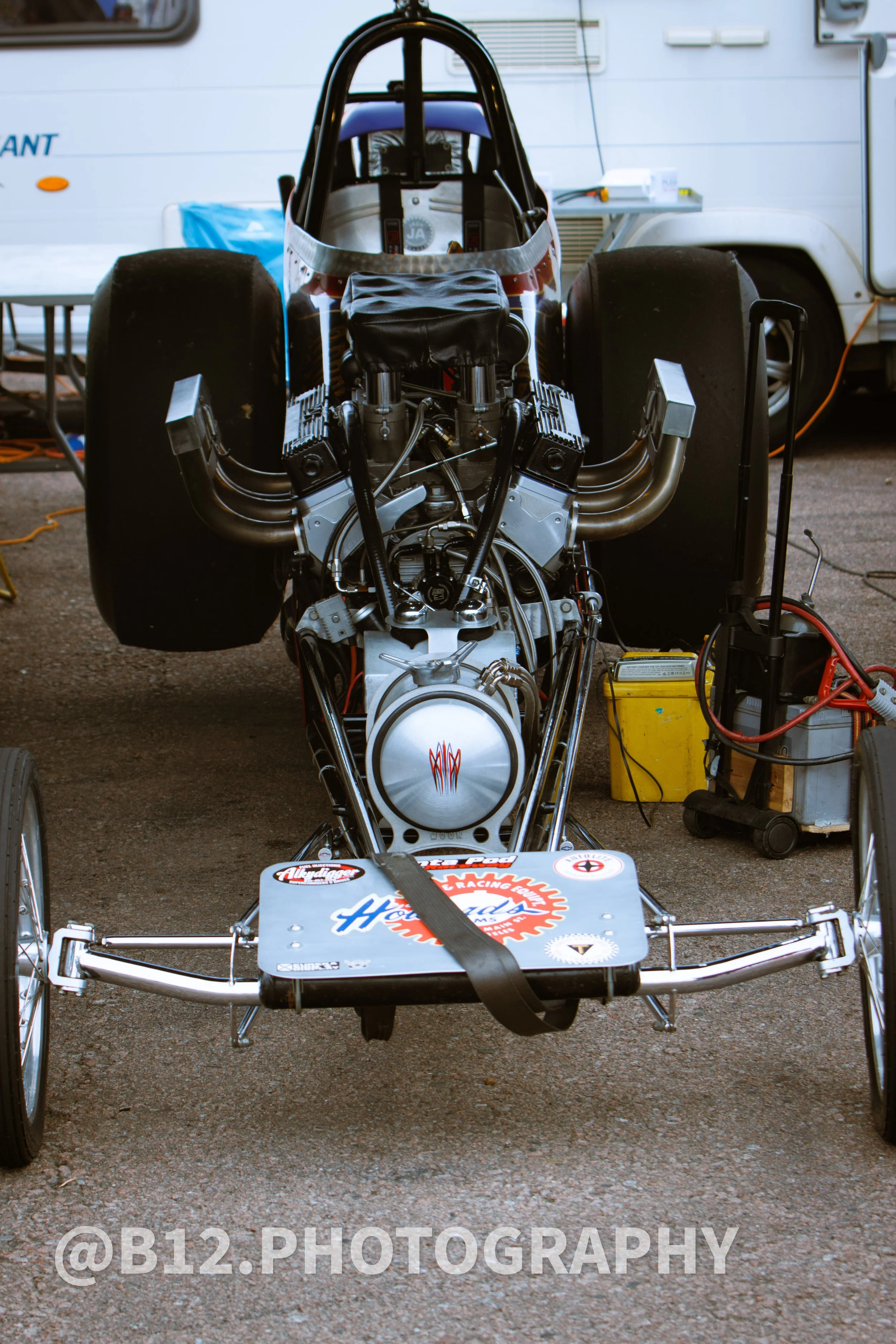 Front view of a vintage race car on a garage floor, showing chassis, engine, and large rear tires, with race stickers on the front platform.