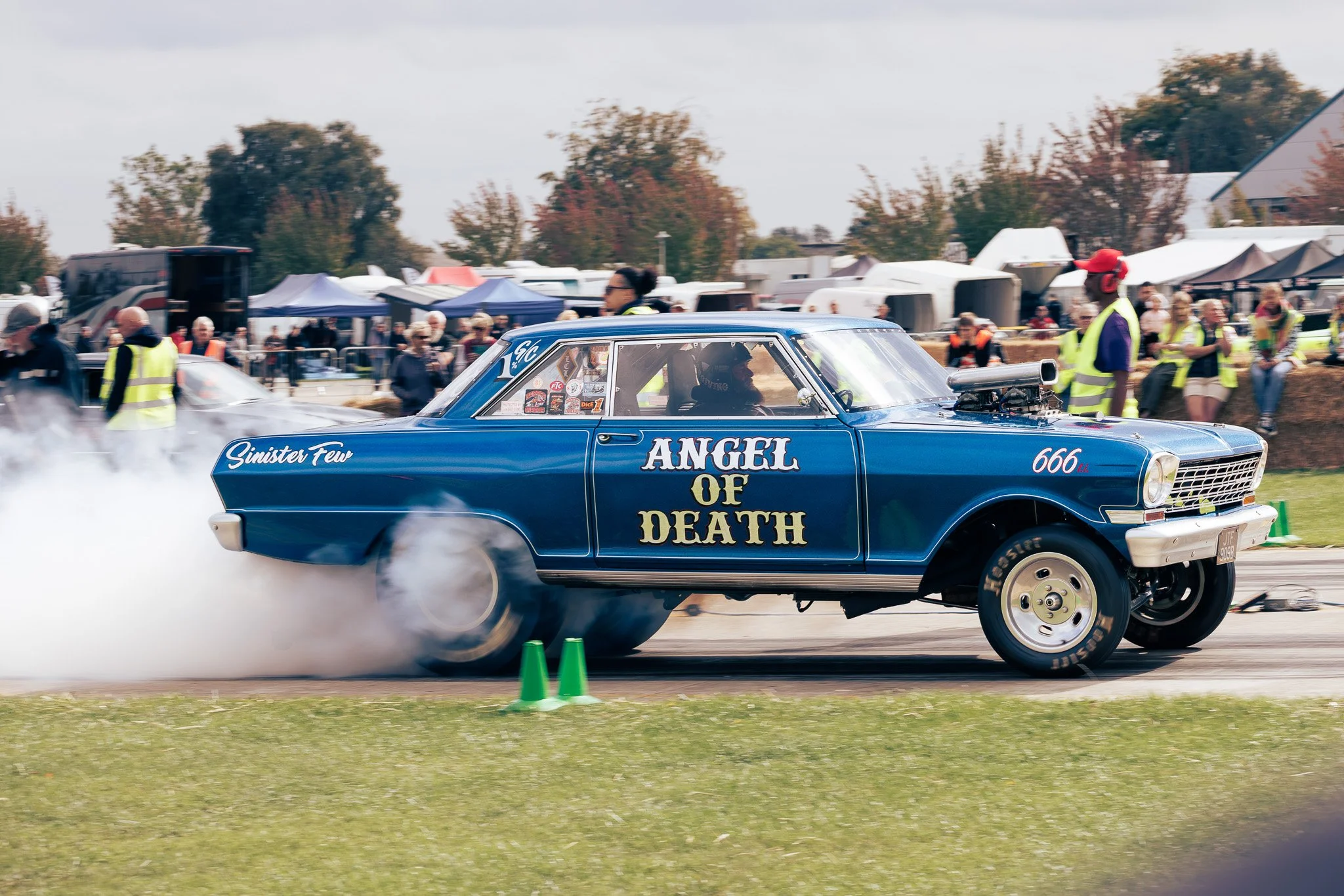 A blue drag racing car with the words 'Angel of Death' on the side, performing a burnout at a race event with spectators and crew in the background.