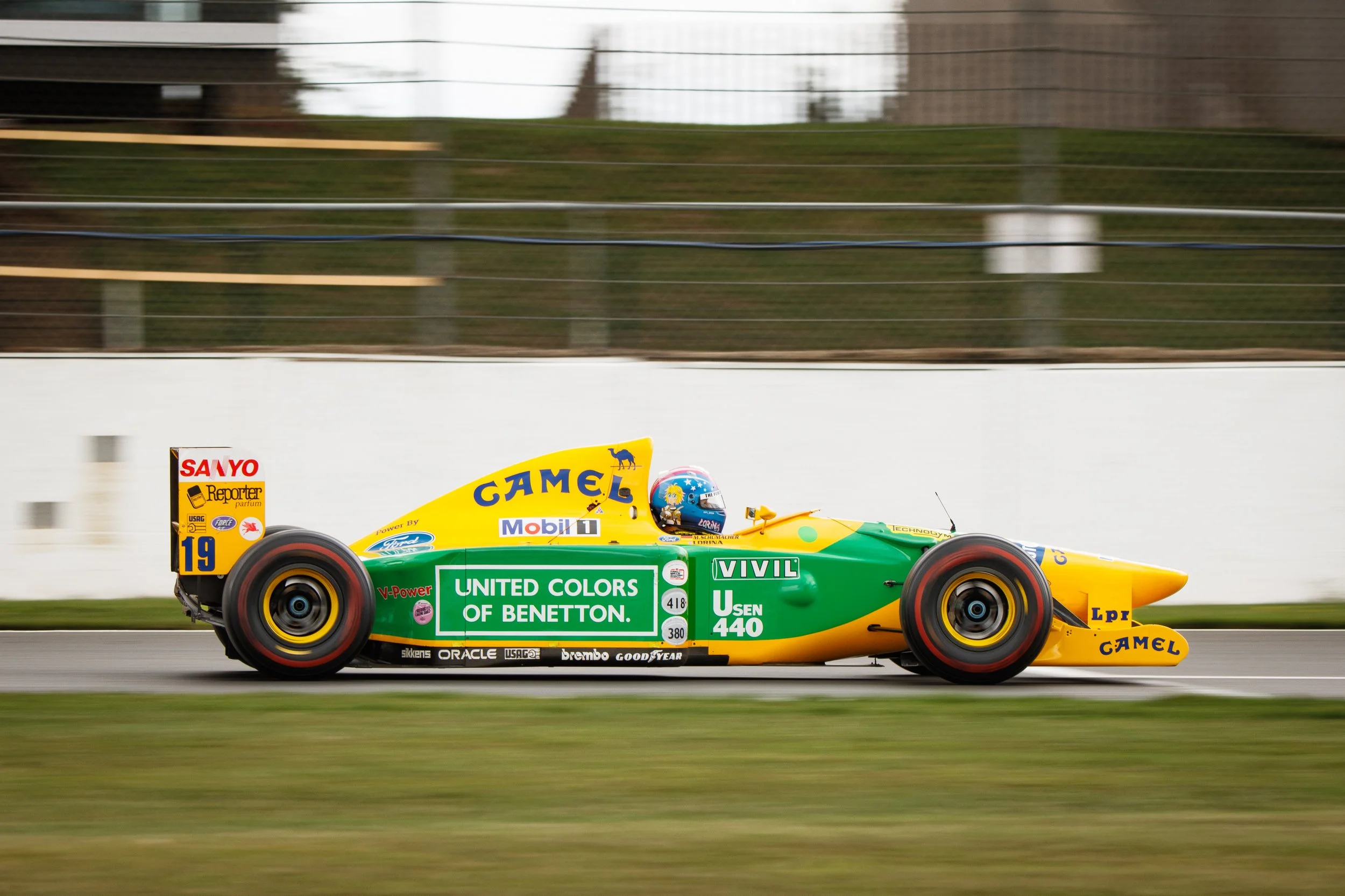 A yellow and green vintage Formula 1 race car speeding on a track, with sponsor logos including Camel, United Colors of Benetton, and Goodyear.