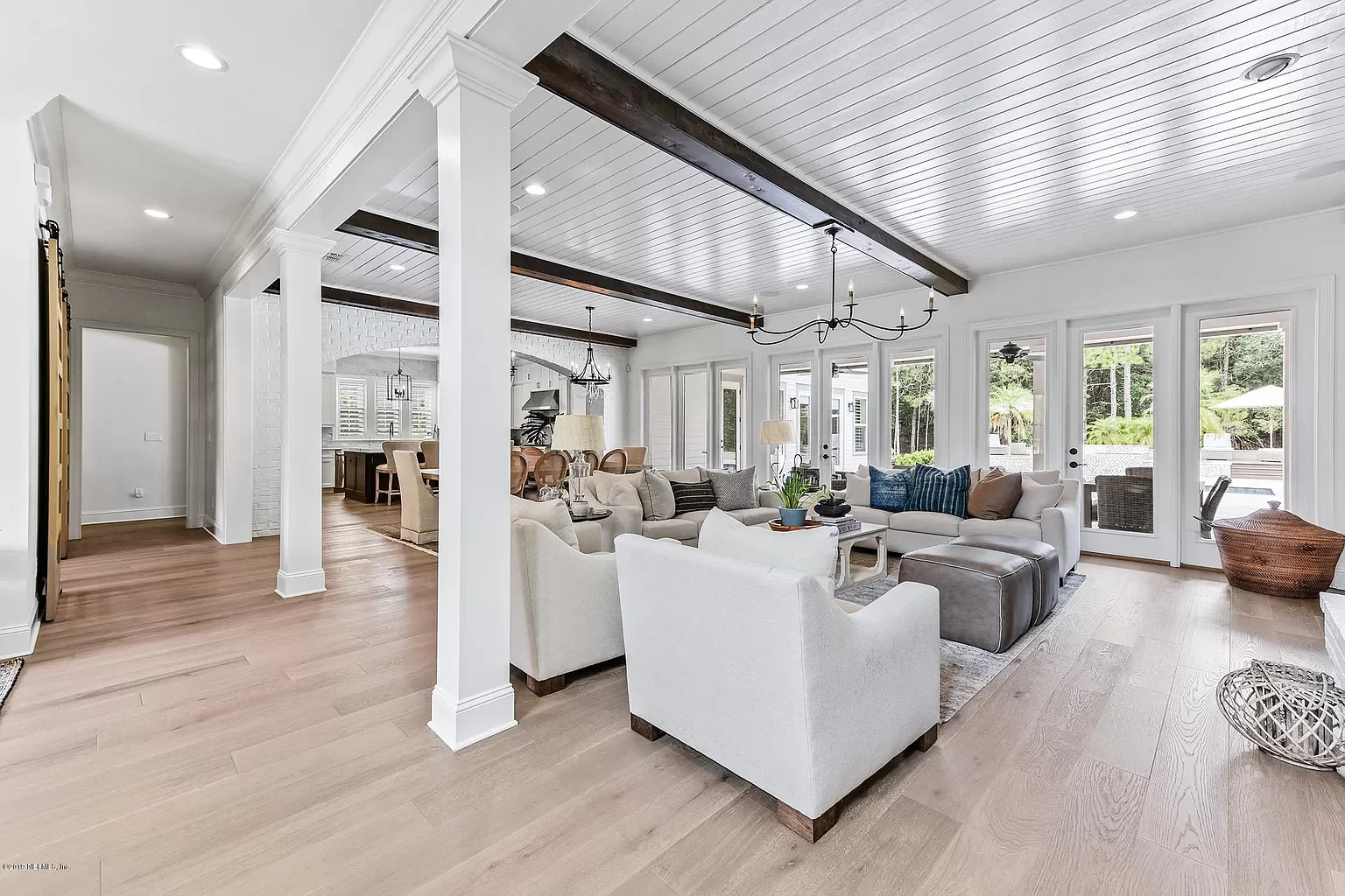 Bright and spacious living room with white sofas, pillows, and large windows showing an outdoor patio with chairs and umbrellas.