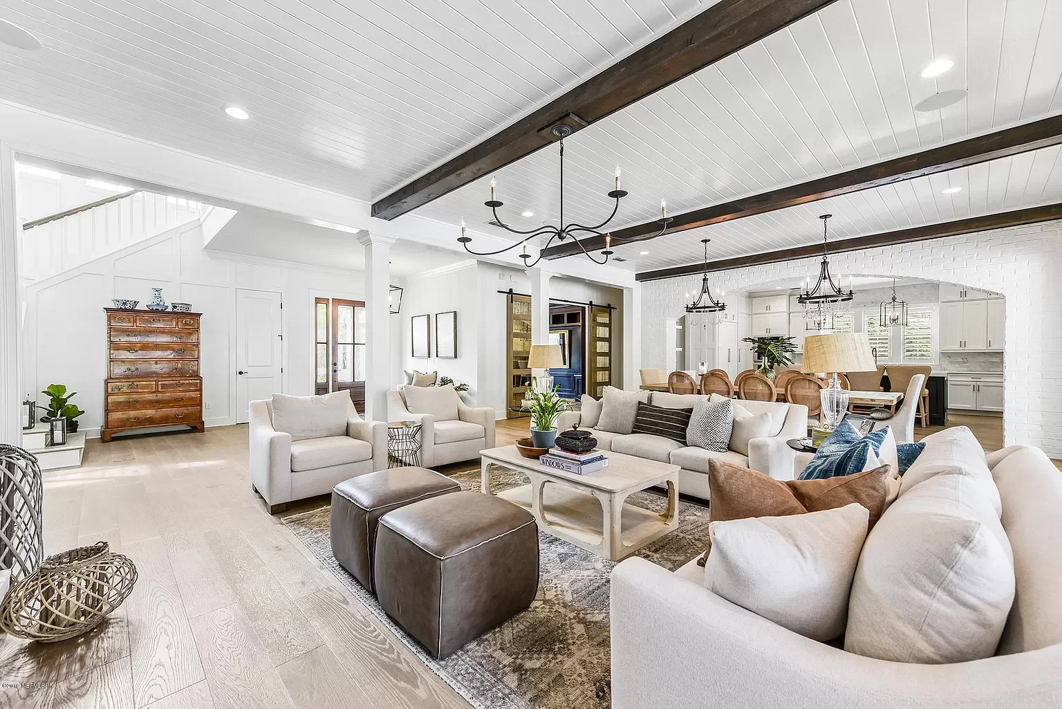 Spacious living room with white walls, hardwood floors, and a white paneled ceiling with dark wooden beams. Includes beige sofas, armchairs, two brown ottomans, a coffee table, and decorative lamps. There is a wooden dresser, potted plants, and a din
