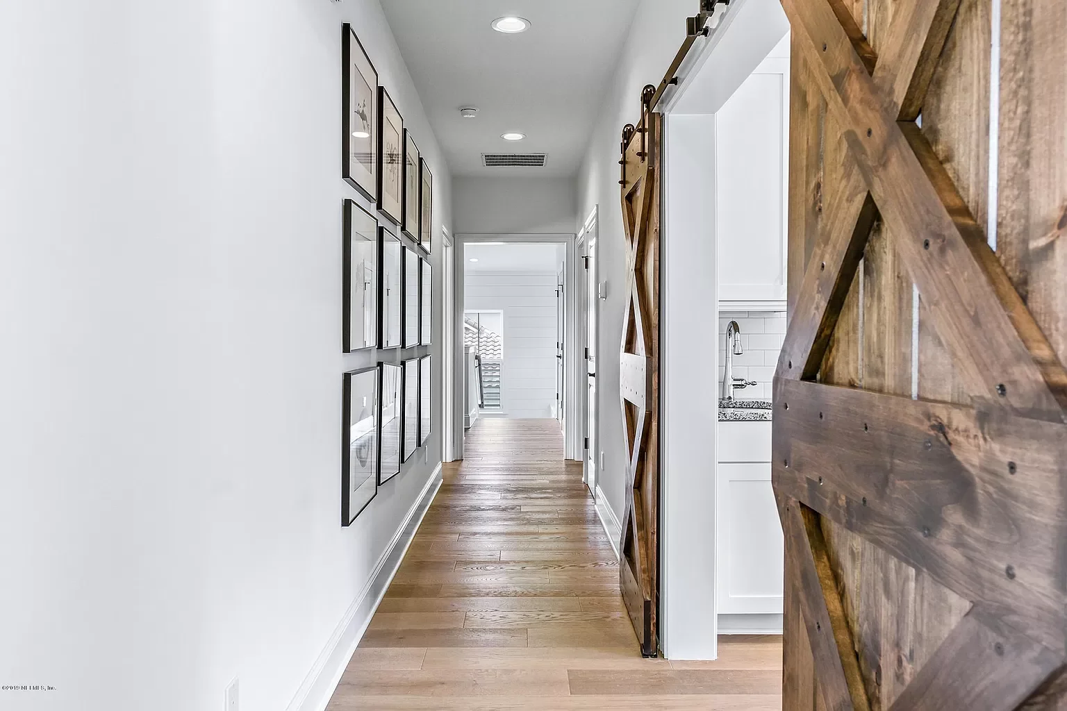 Interior view of a hallway with white walls, wooden flooring, framed pictures on the left wall, and a sliding barn door on the right side leading to a kitchen area.
