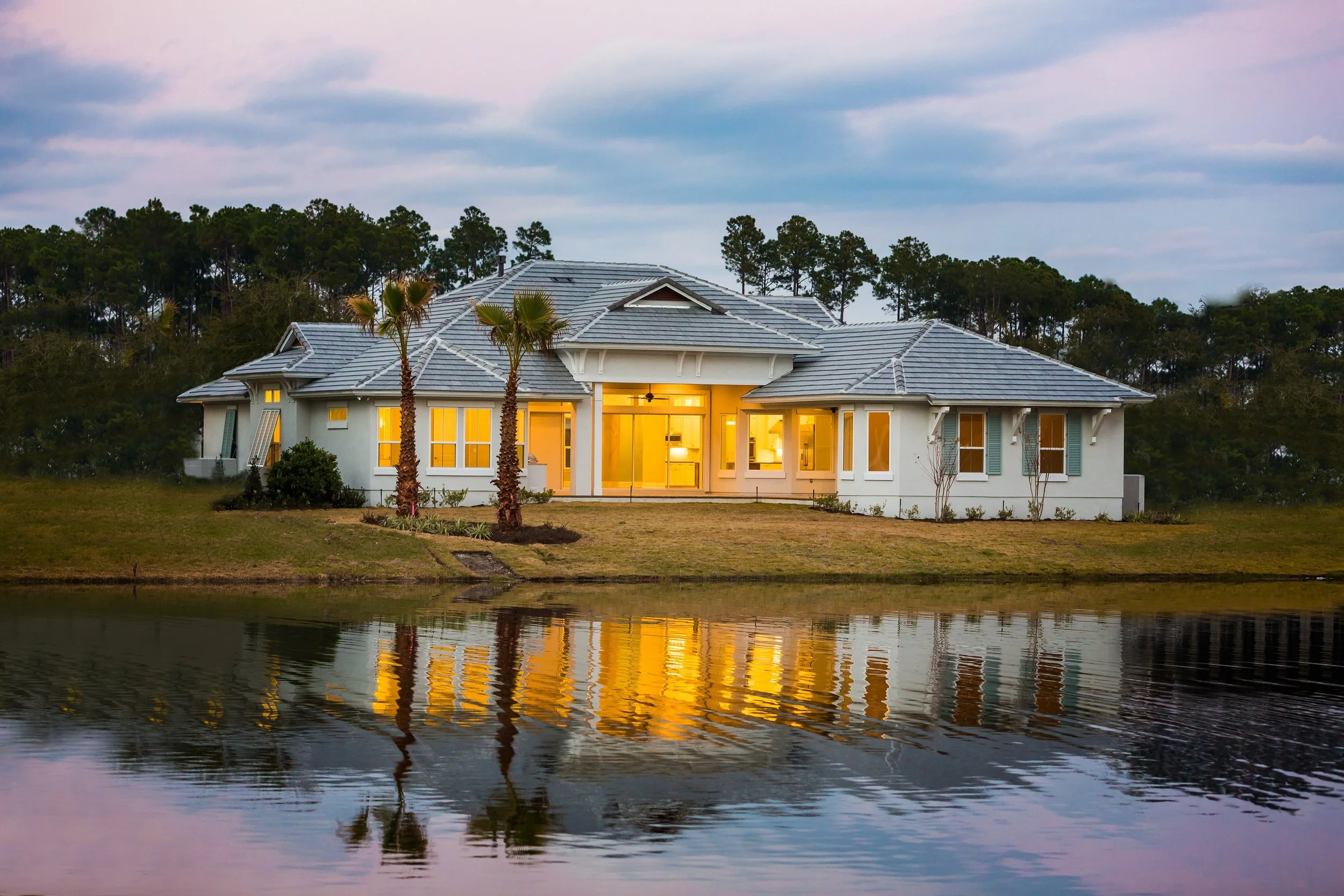 A large, modern house with white walls and a grey tiled roof, illuminated from within, situated on a grassy bank overlooking a calm body of water with reflections of the house, trees, and sky during dusk.