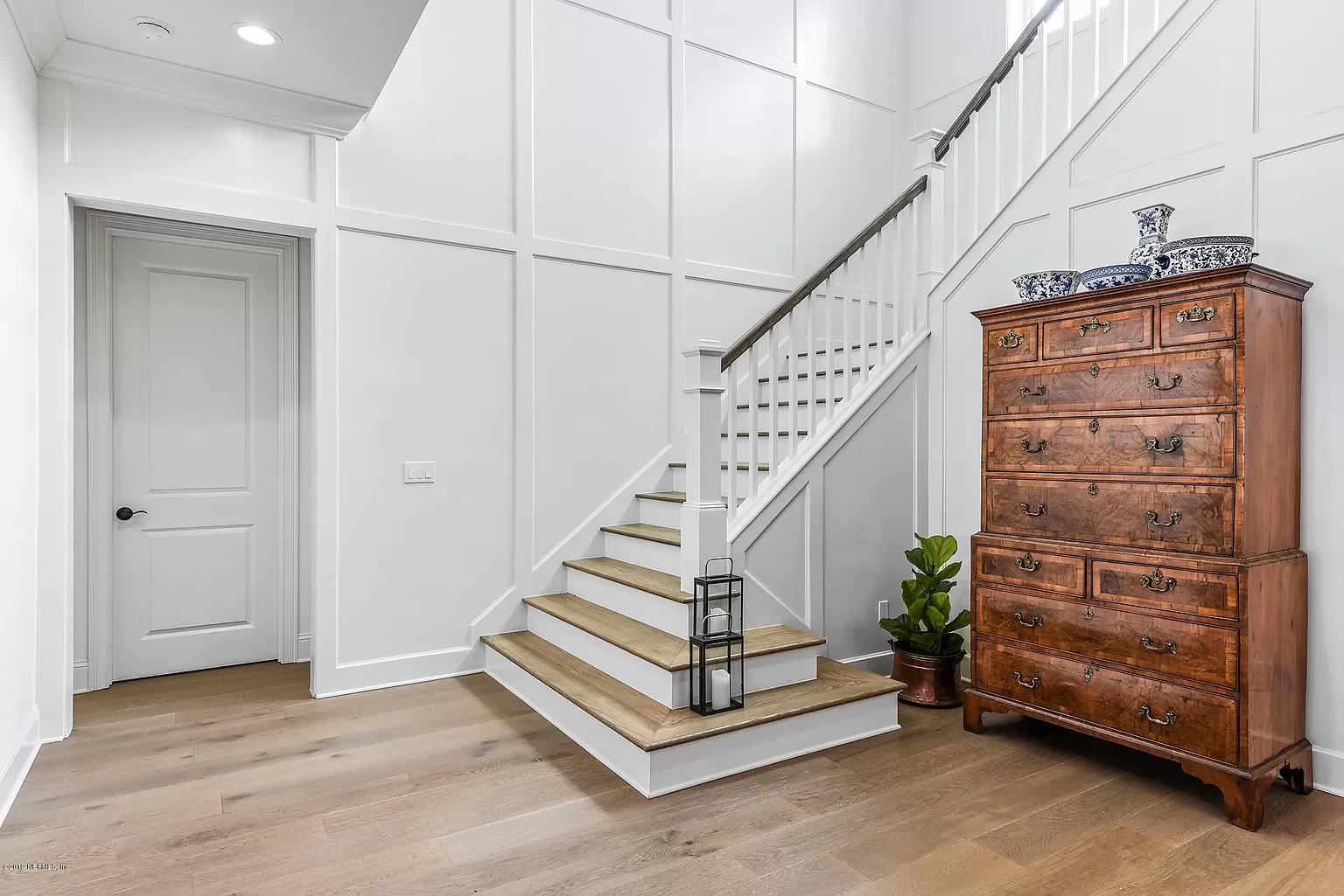 Interior of a modern home with white walls, wooden flooring, a staircase with wooden steps and white risers, a closed door, a wooden chest of drawers, a potted plant, and decorative blue and white ceramics on top of the chest.