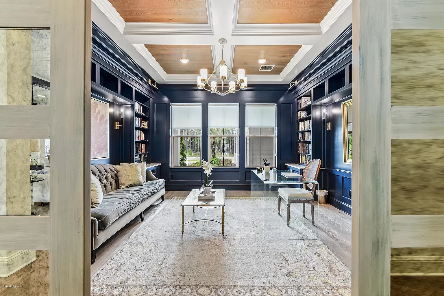 Living room with navy blue walls, white trim, large windows, bookshelf, cream sofa, glass coffee table, armchair, light-colored rug, chandelier, and decorative items.