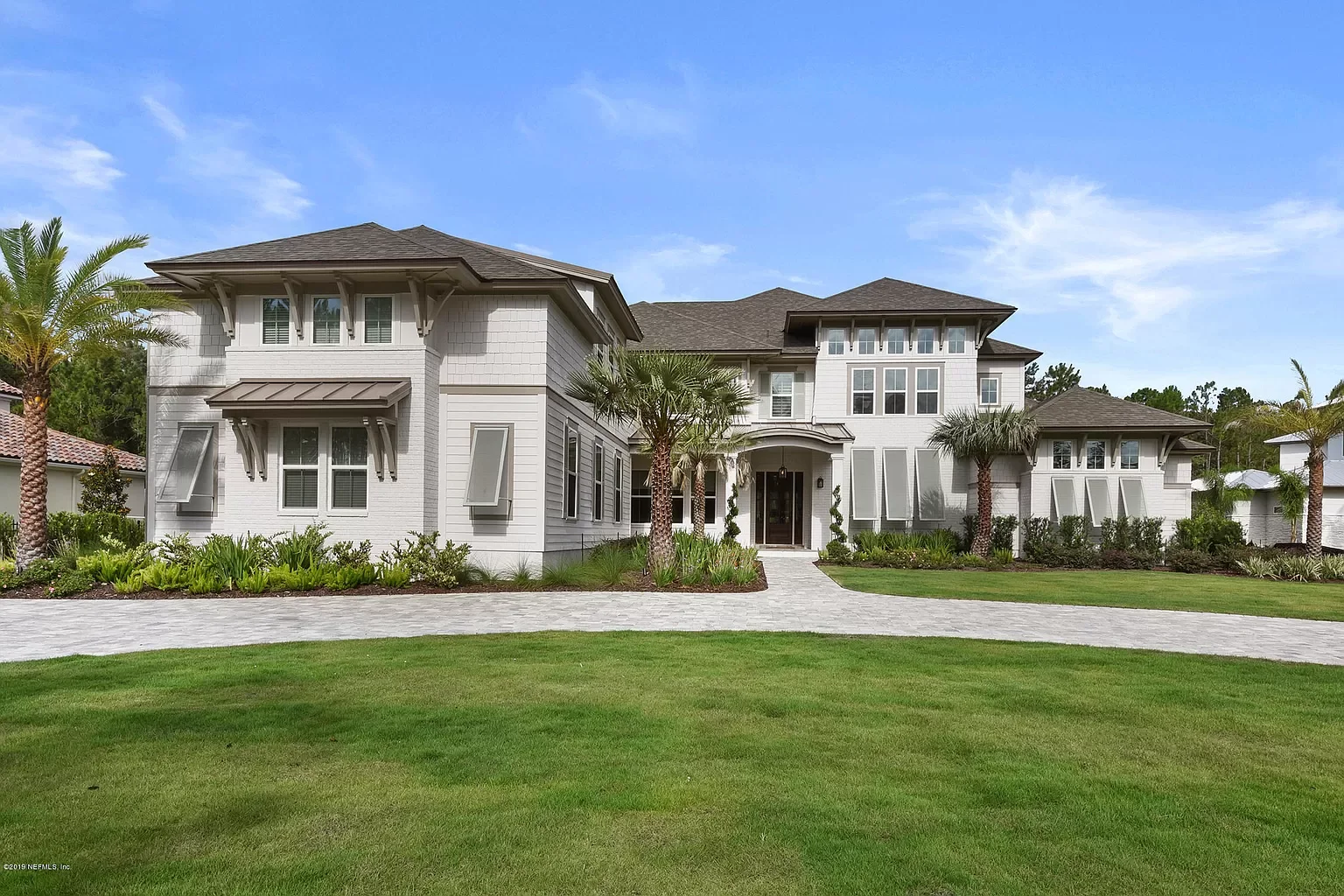 A large, white, two-story house with multiple windows, a front porch, and a curved walkway leading to the entrance. The house is surrounded by a well-maintained green lawn and palm trees.