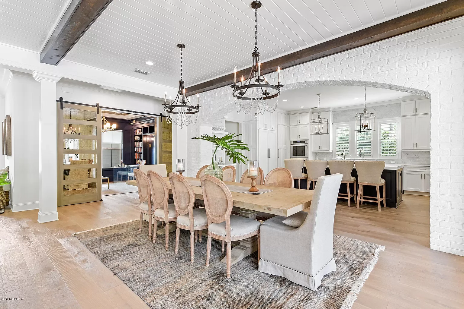 Bright open-concept dining and kitchen area with white walls, wood beams, and hardwood floors, featuring a large dining table with eight chairs, a rug, and modern light fixtures.