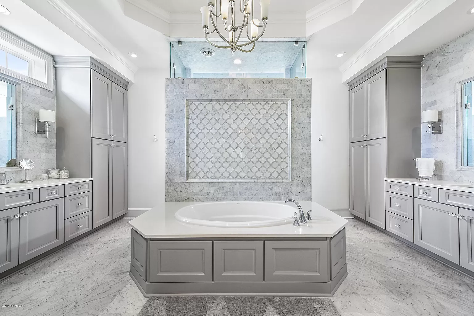 A spacious and luxurious bathroom with a large bathtub in the center, surrounded by light gray cabinetry, marble flooring, and walls. There are two windows, a chandelier overhead, and a glass-walled shower at the back.