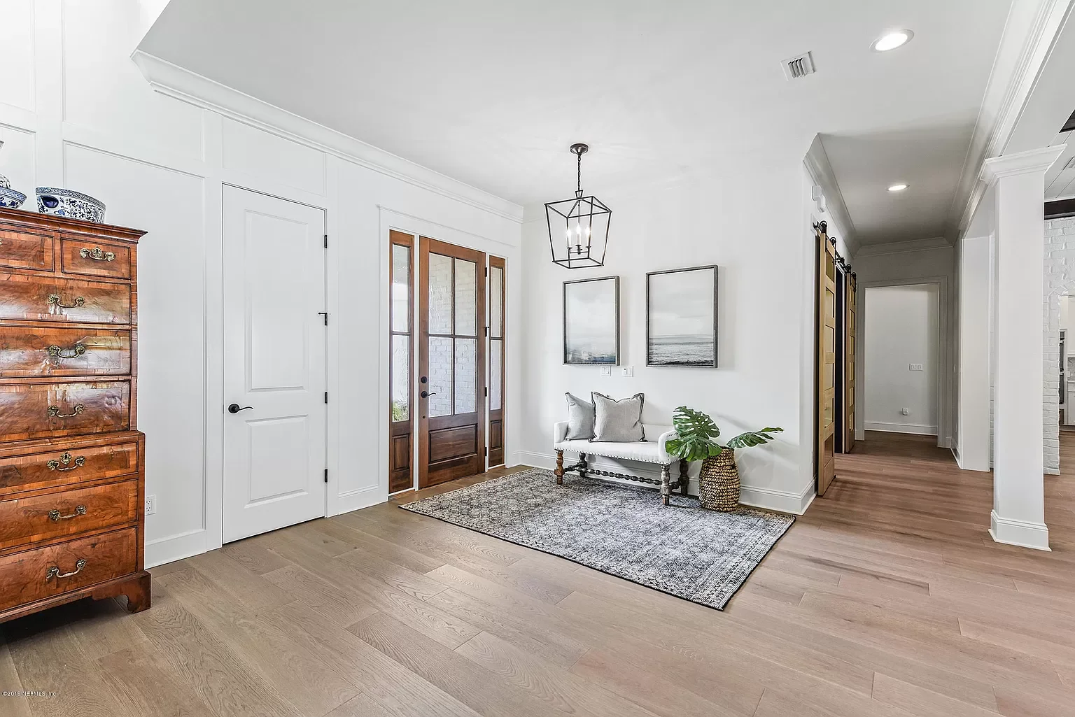 Entrance area of a home with a wooden door, white walls, a small white bench with pillows, a potted plant, artwork on the wall, and a wooden dresser.