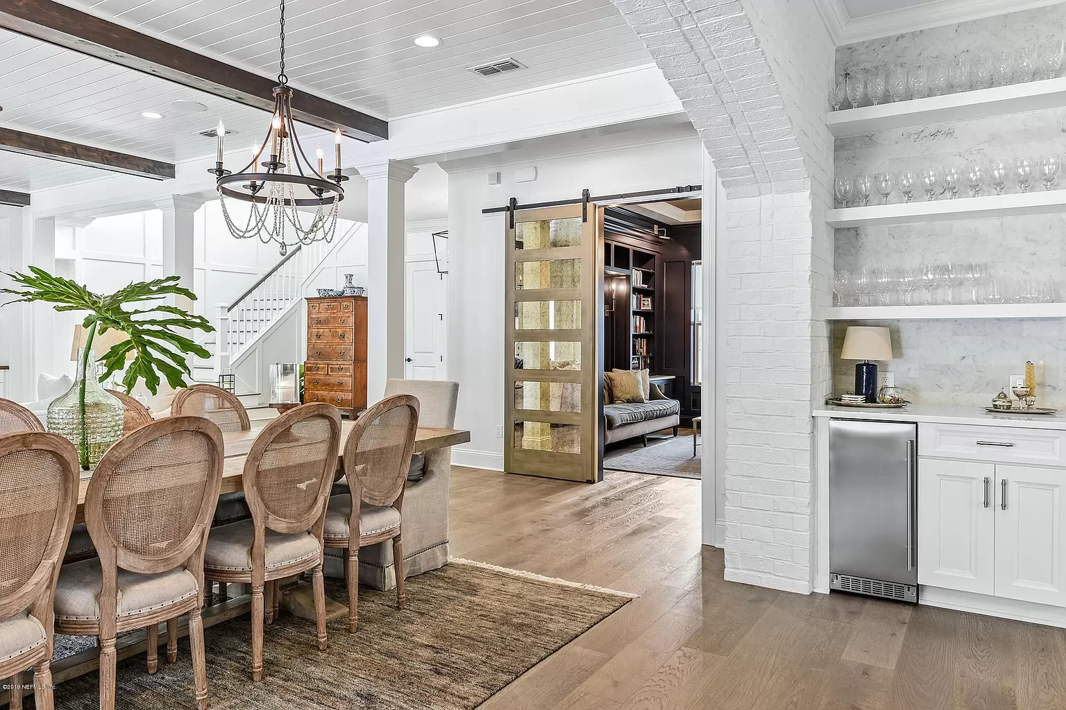 Interior view of a dining and living area with a white brick wall, a sliding barn door, wooden dining table with six chairs, chandelier, and a nook with shelves and decorative items.
