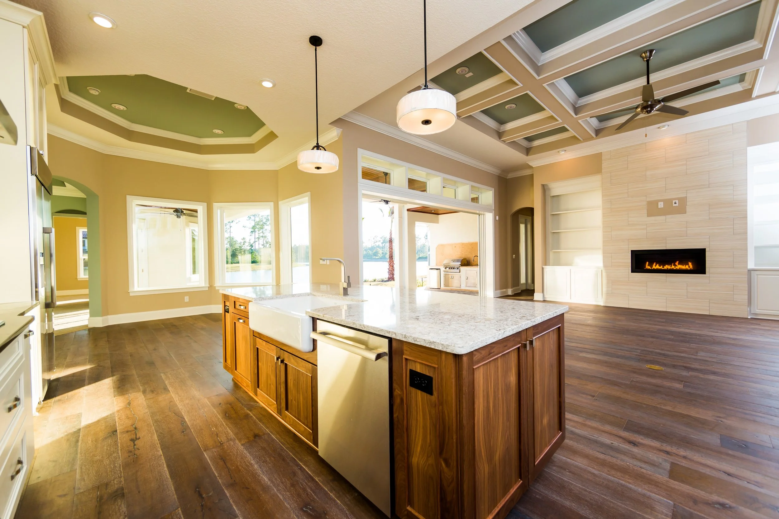 Open-concept kitchen and living room with hardwood floors, kitchen island, built-in shelves, gas fireplace, large windows, and a ceiling with coffered detail.