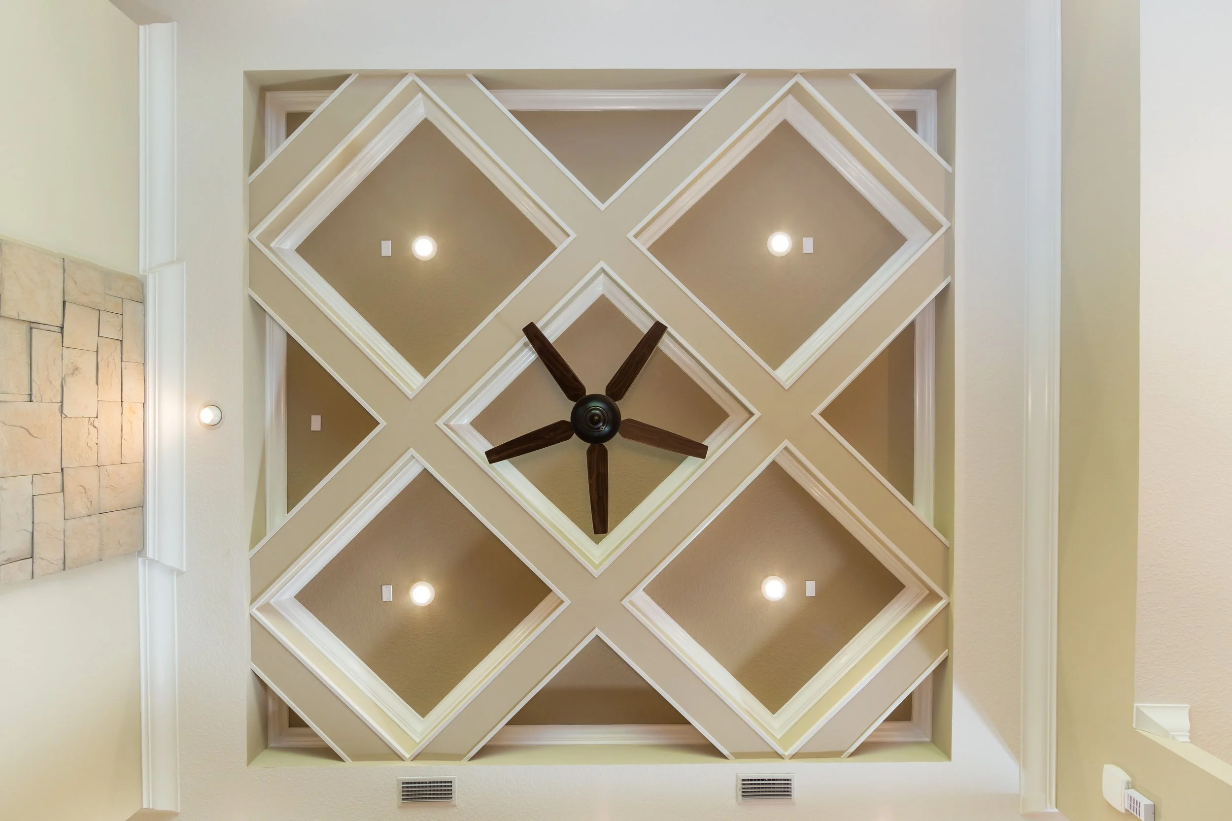 Ceiling with a large, decorative geometric design featuring diamond shapes, with recessed lighting and a central ceiling fan with five dark wooden blades.