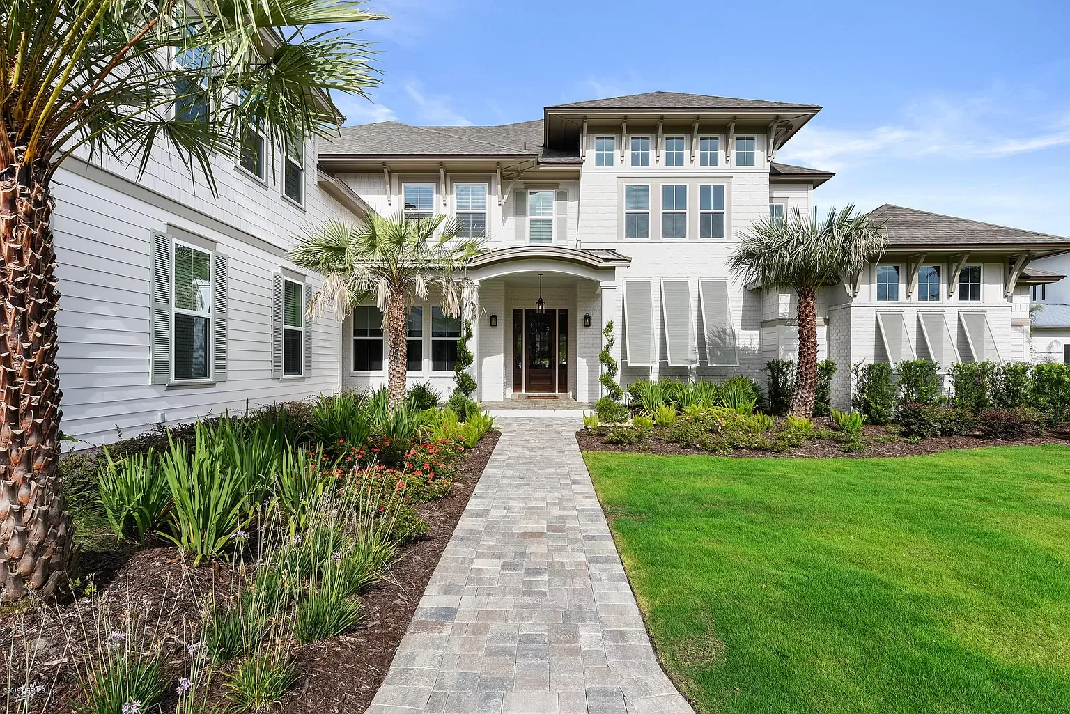 Front view of a large white house with a paved walkway leading to the front door, surrounded by a well-maintained lawn and tropical plants, including palm trees.