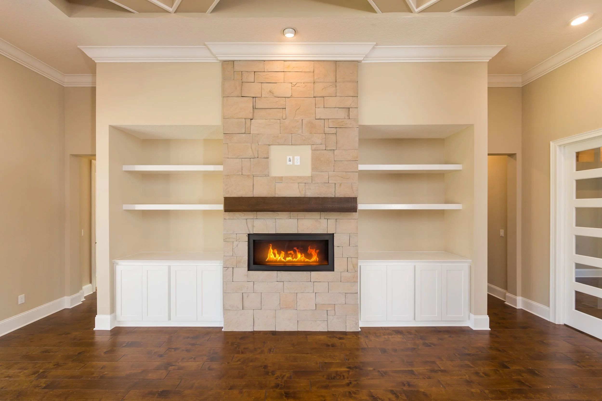 Living room with a built-in fireplace set in a stone wall with wooden mantel, flanked by white shelves and cabinets, hardwood floor, and beige walls.