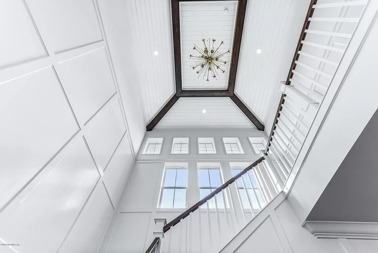 Interior view looking upwards at a staircase and high windows, with a ceiling featuring a chandelier and dark wood trim.