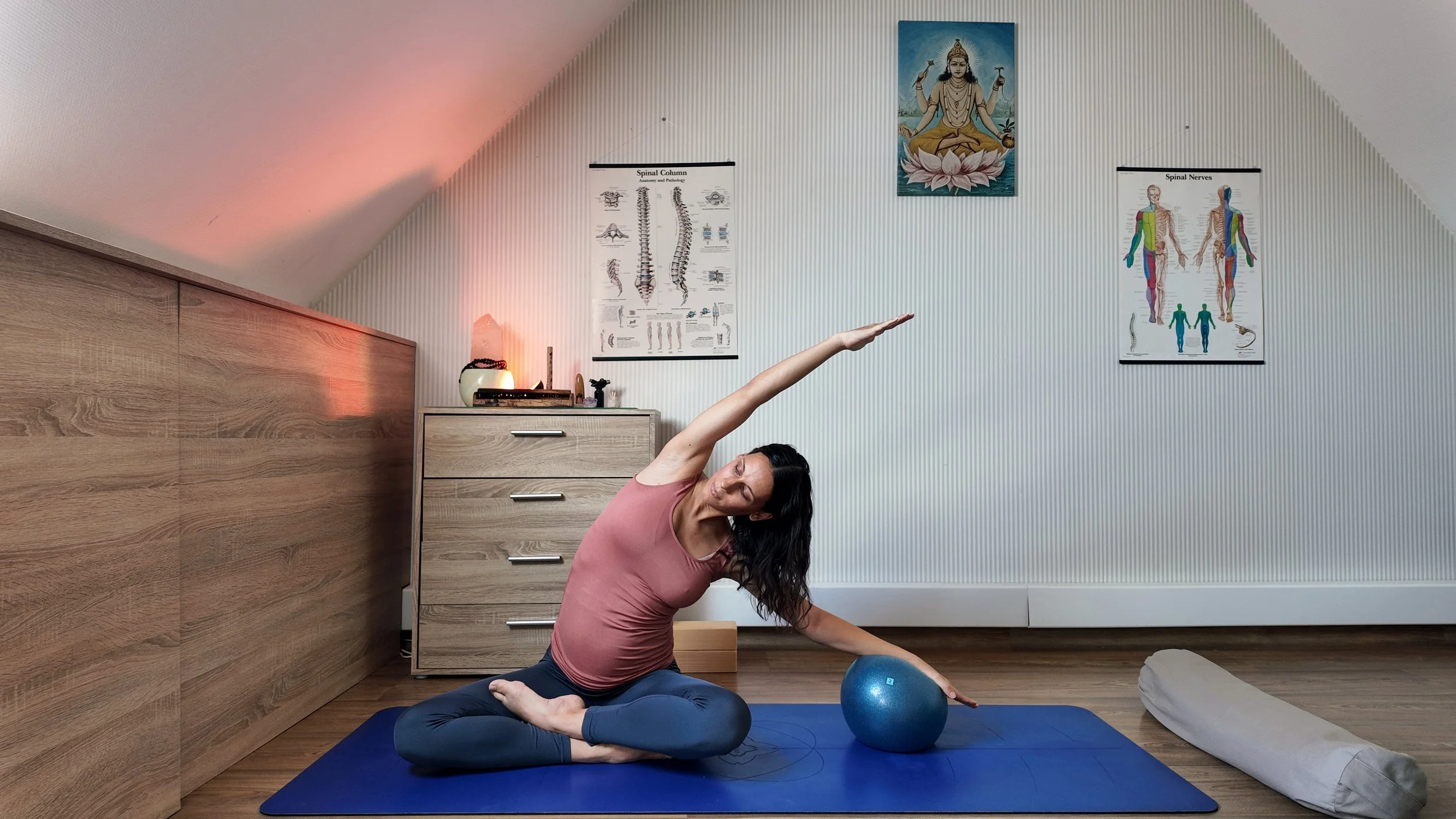 A woman practicing yoga indoors, seated on a blue yoga mat, performing a side stretch with her right arm extended overhead. She is wearing a sleeveless pink top and dark leggings. The room has wooden floors, a white and wood-paneled wall, and yoga posters, with a painting above her.