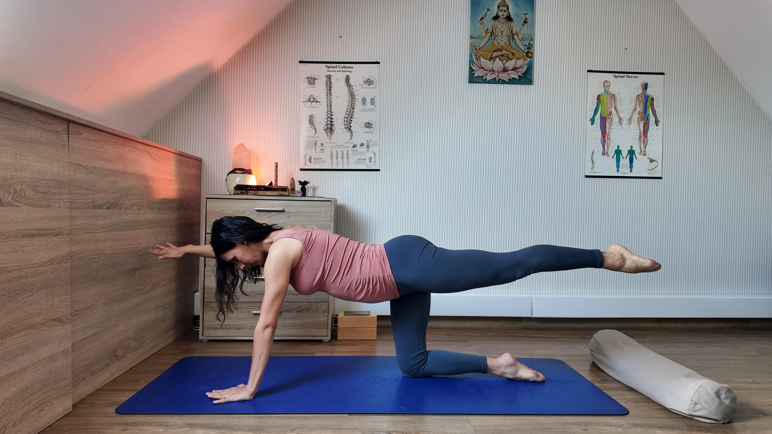 A woman practicing yoga in a room with spiritual posters and a painting of a deity on the wall. She is in a balancing pose with one leg extended back and her hands in a prayer position against the wall.