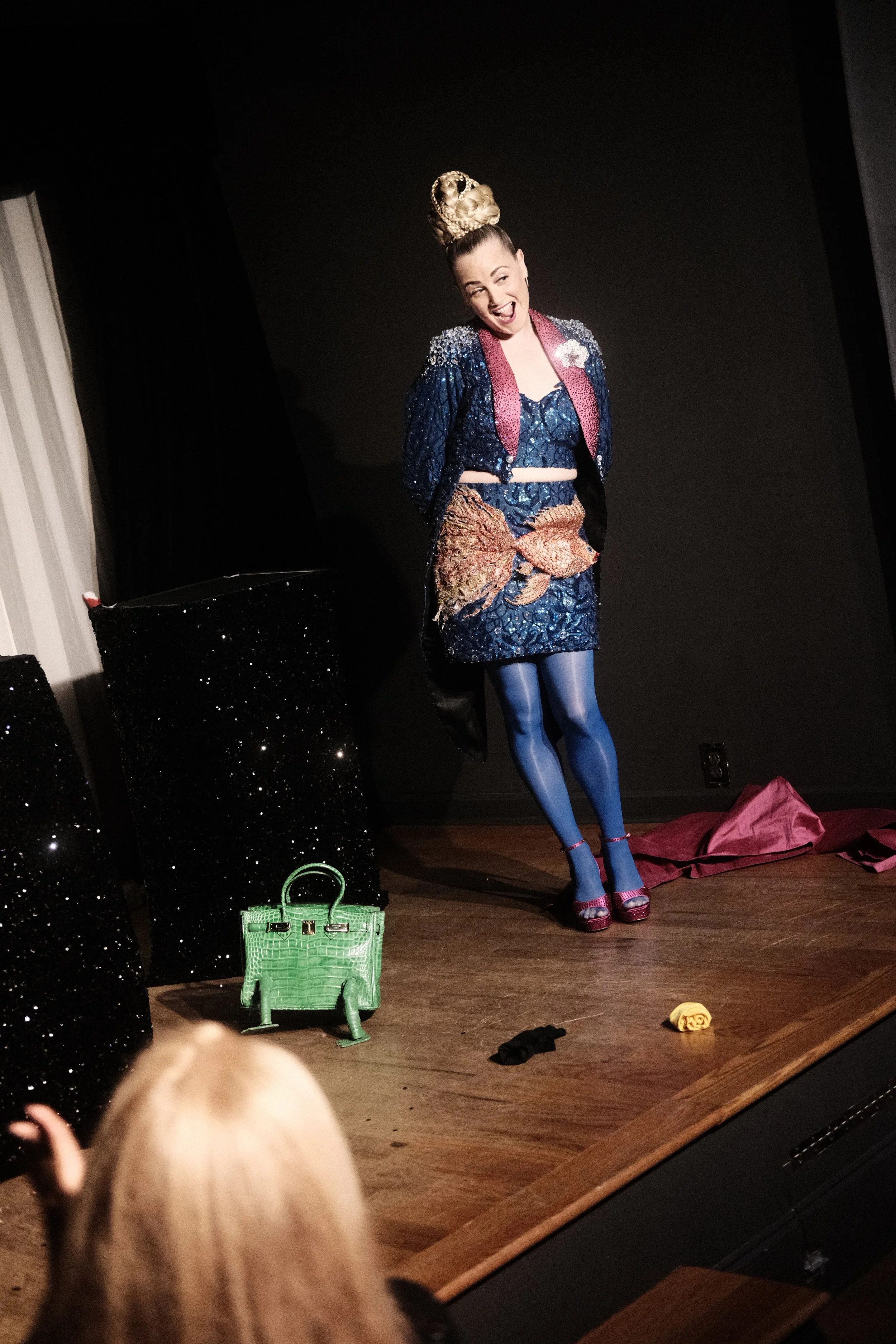 Performer on stage wearing a colorful outfit, with a large ornate hairpiece, blue tights, and high heels, standing against a black background with props and audience visible.