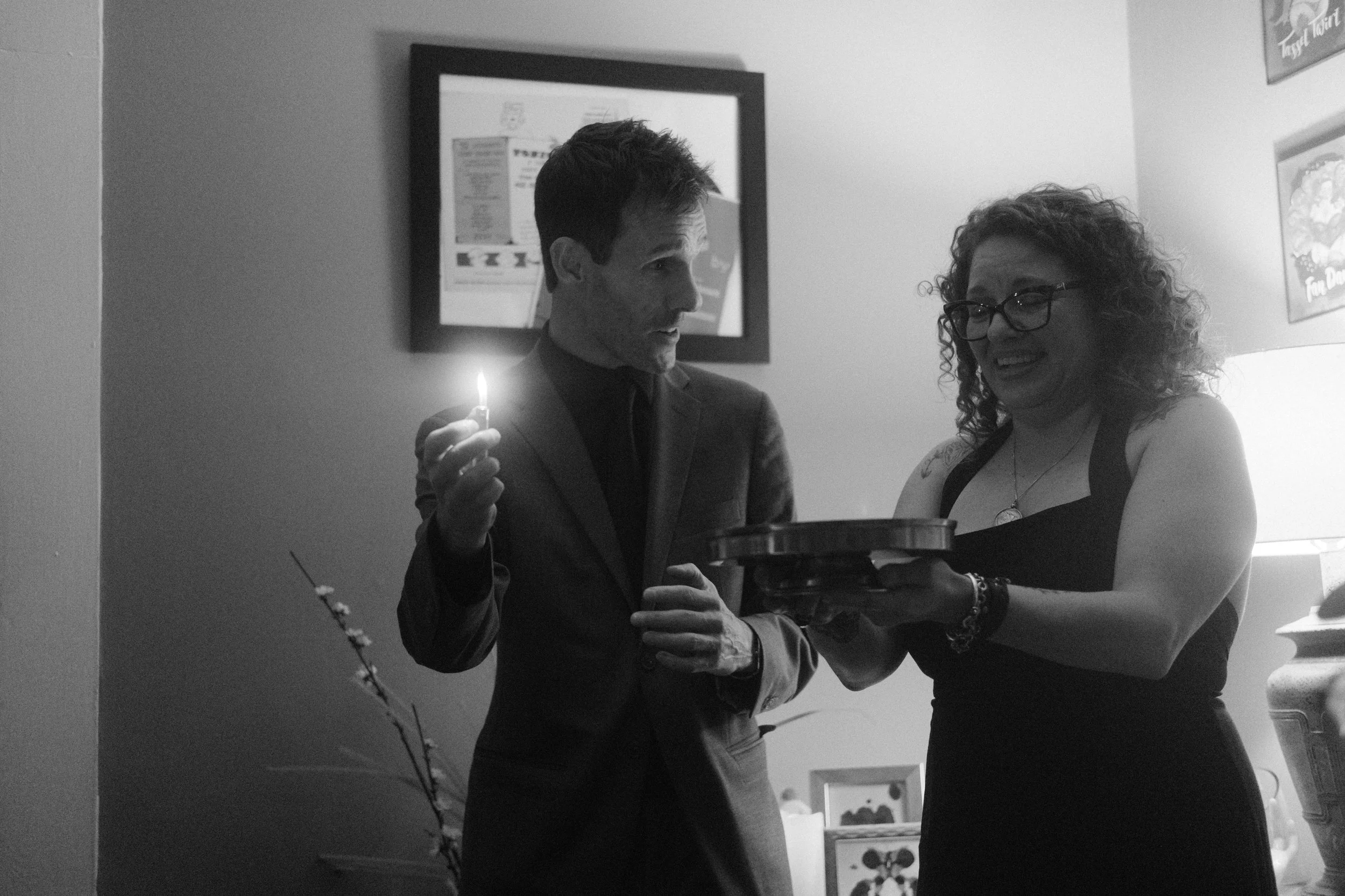 A man in a suit holding a small lit candle and a woman with curly hair and glasses smiling and serving a plate, in a dimly lit room.