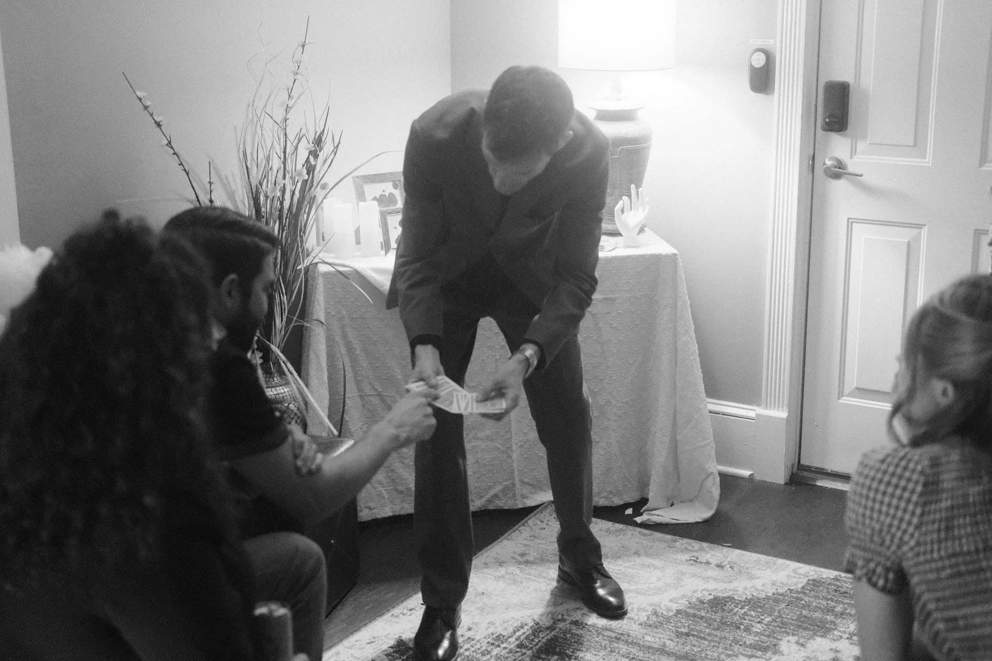 A man in a suit is handing money to a woman seated on a chair, with two other women seated nearby in a room with decorated walls and a table with candles, a framed picture, and decorative items.