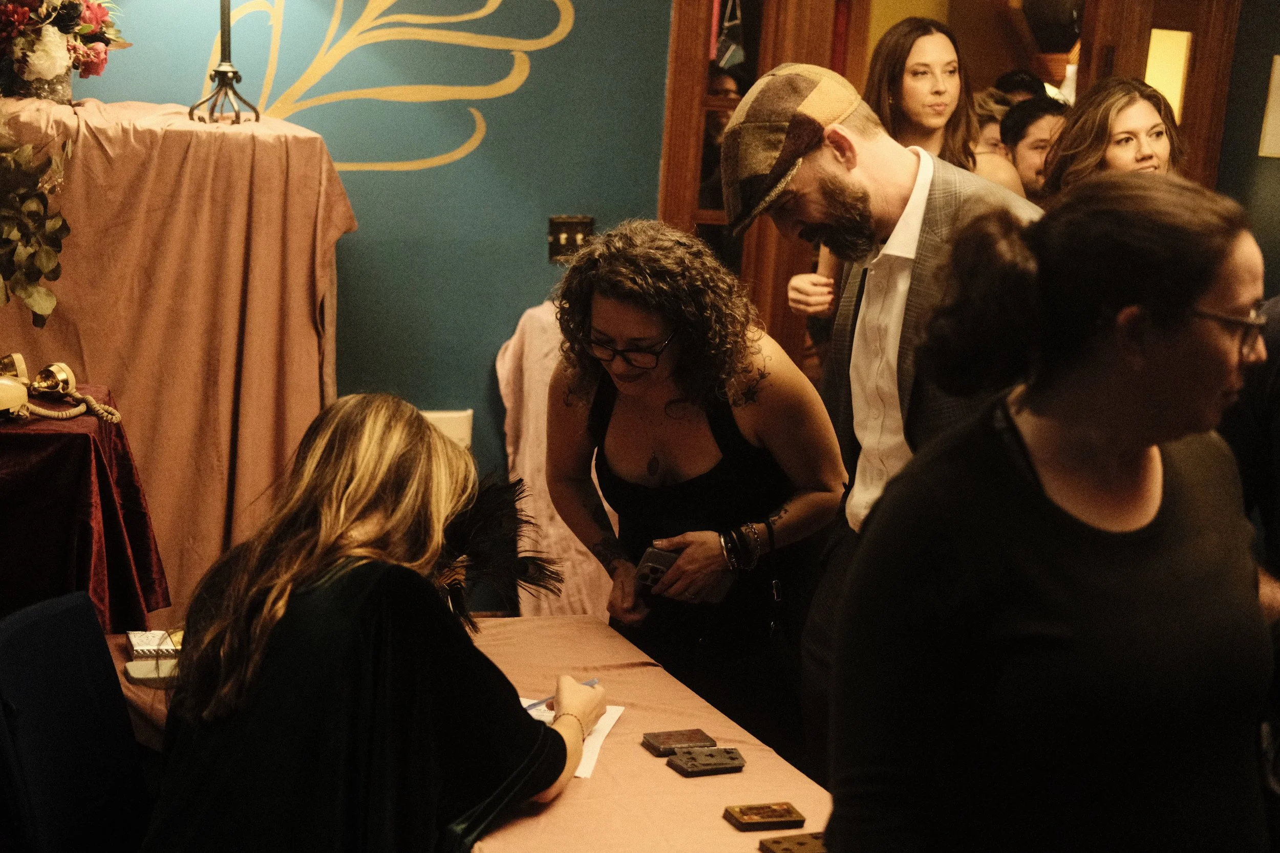 Group of people gathered around a table at a social event, with a woman signing an item and others observing.