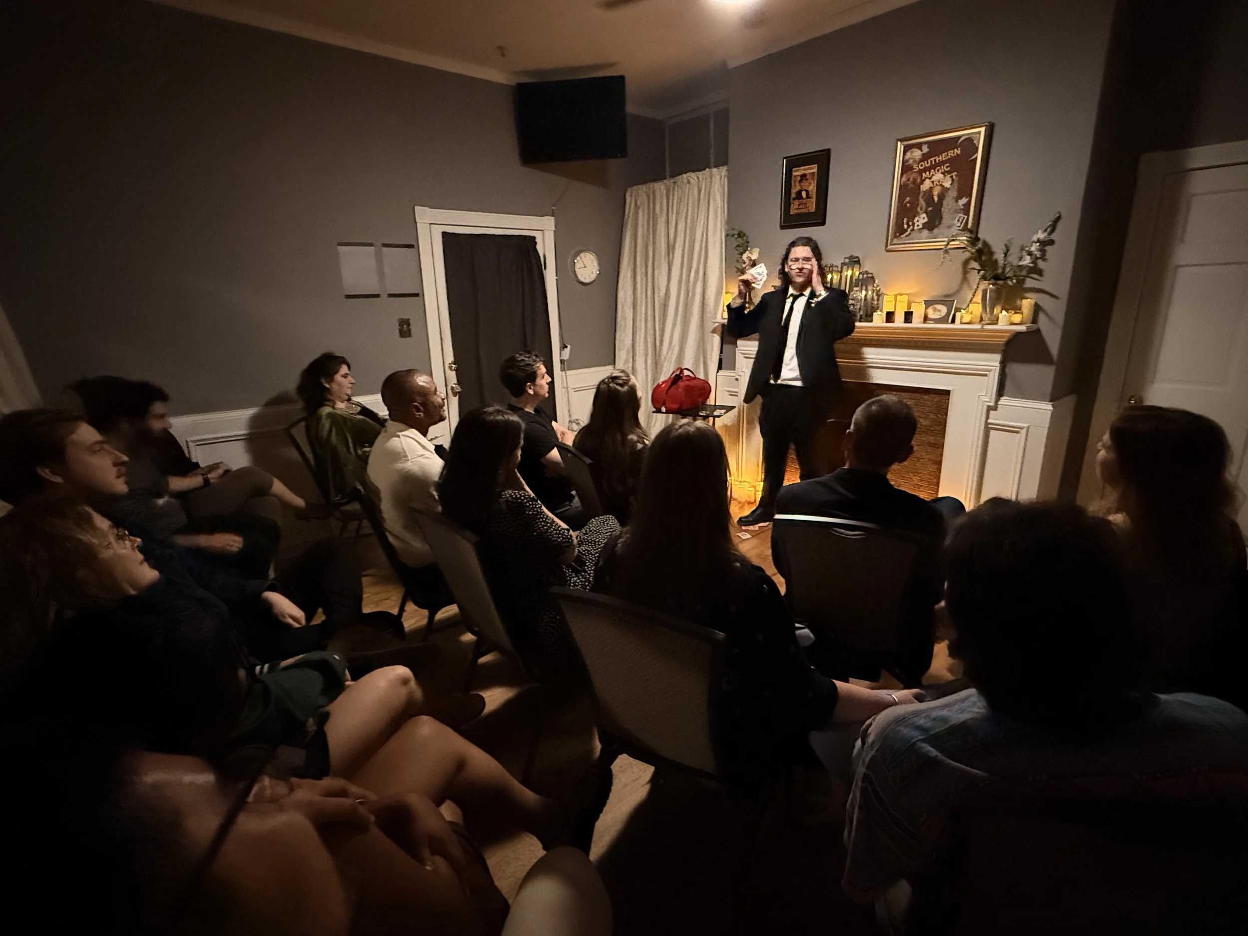 A man in a suit standing in front of an audience, holding a bottle and a glass, speaking or performing in a cozy, dimly lit room with a fireplace, framed artwork, and decorative candles.