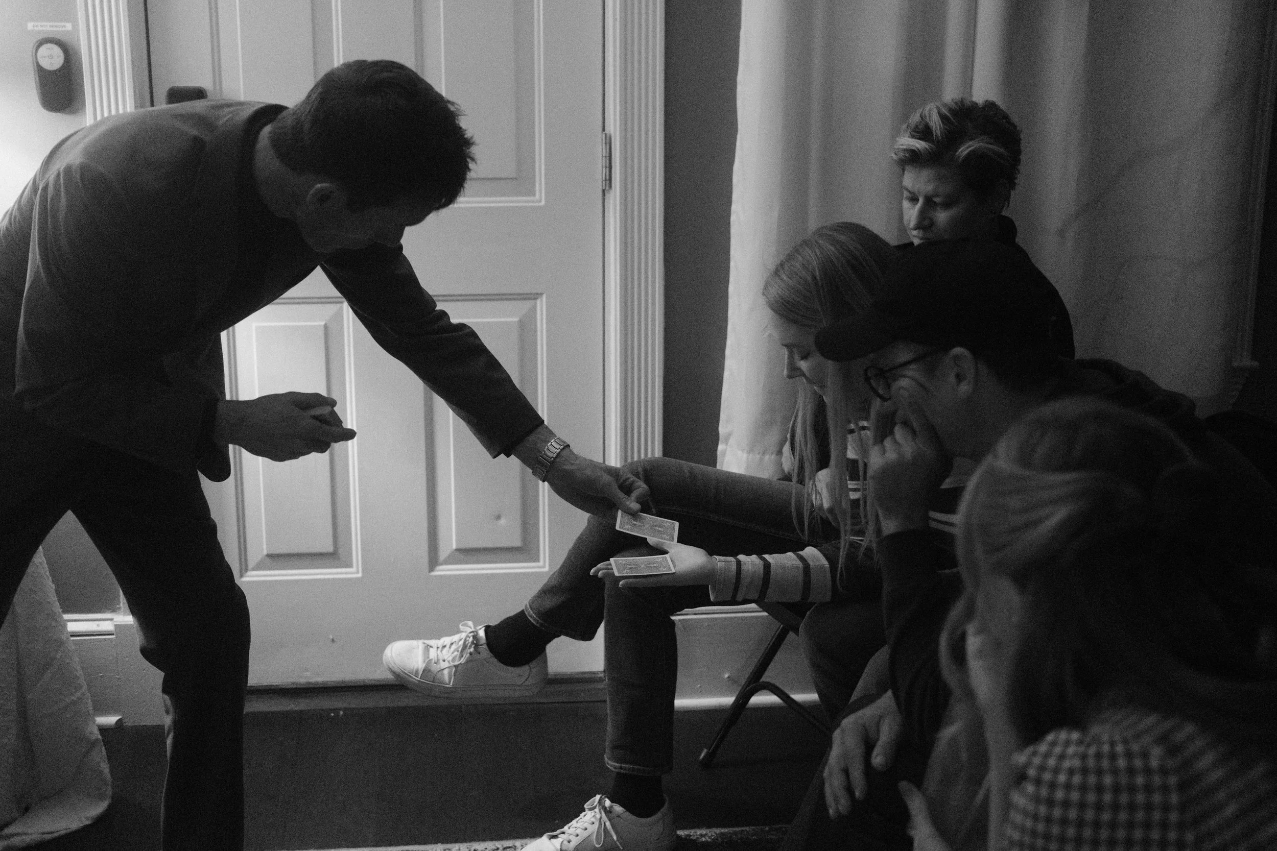 A man is performing a magic card trick for four attentive people sitting on a bench indoors.