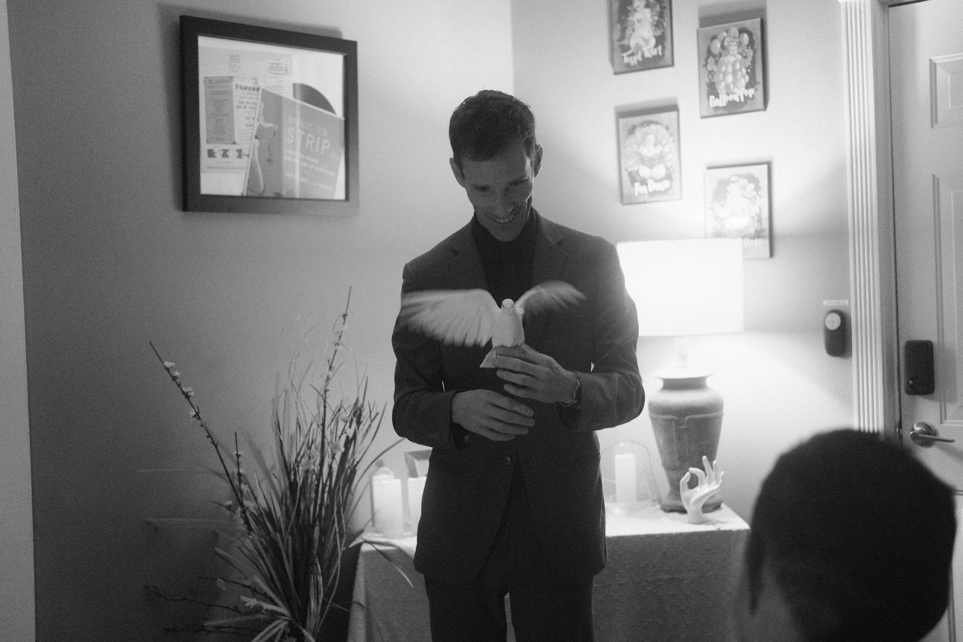 A man in a suit holding a white pigeon in a living room with artwork on the wall, a lamp, and decorative items.