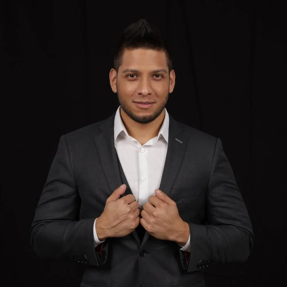 A young man in a dark suit and white shirt adjusting his suit jacket against a black background.