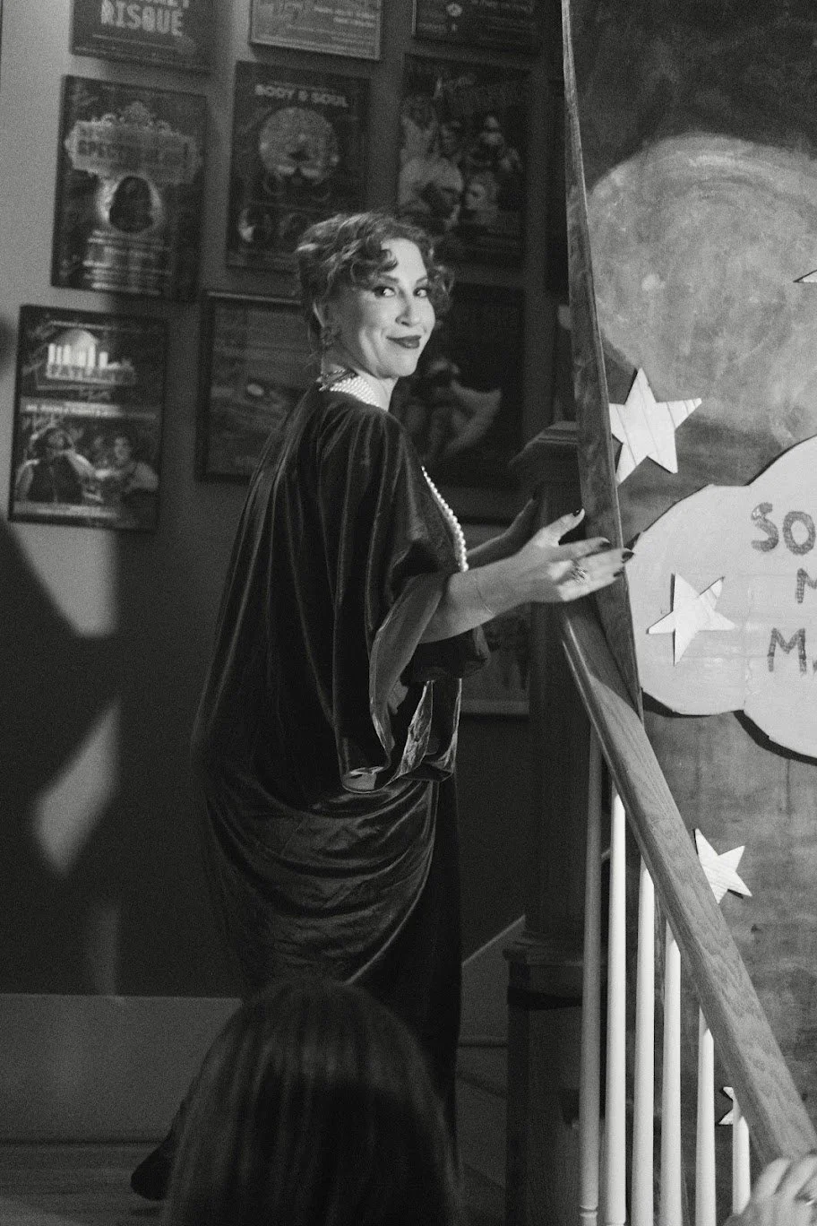 A woman with short, curly hair, wearing a velvet dress and pearl necklace, smiling and standing near an easel with stars, in a room decorated with posters.