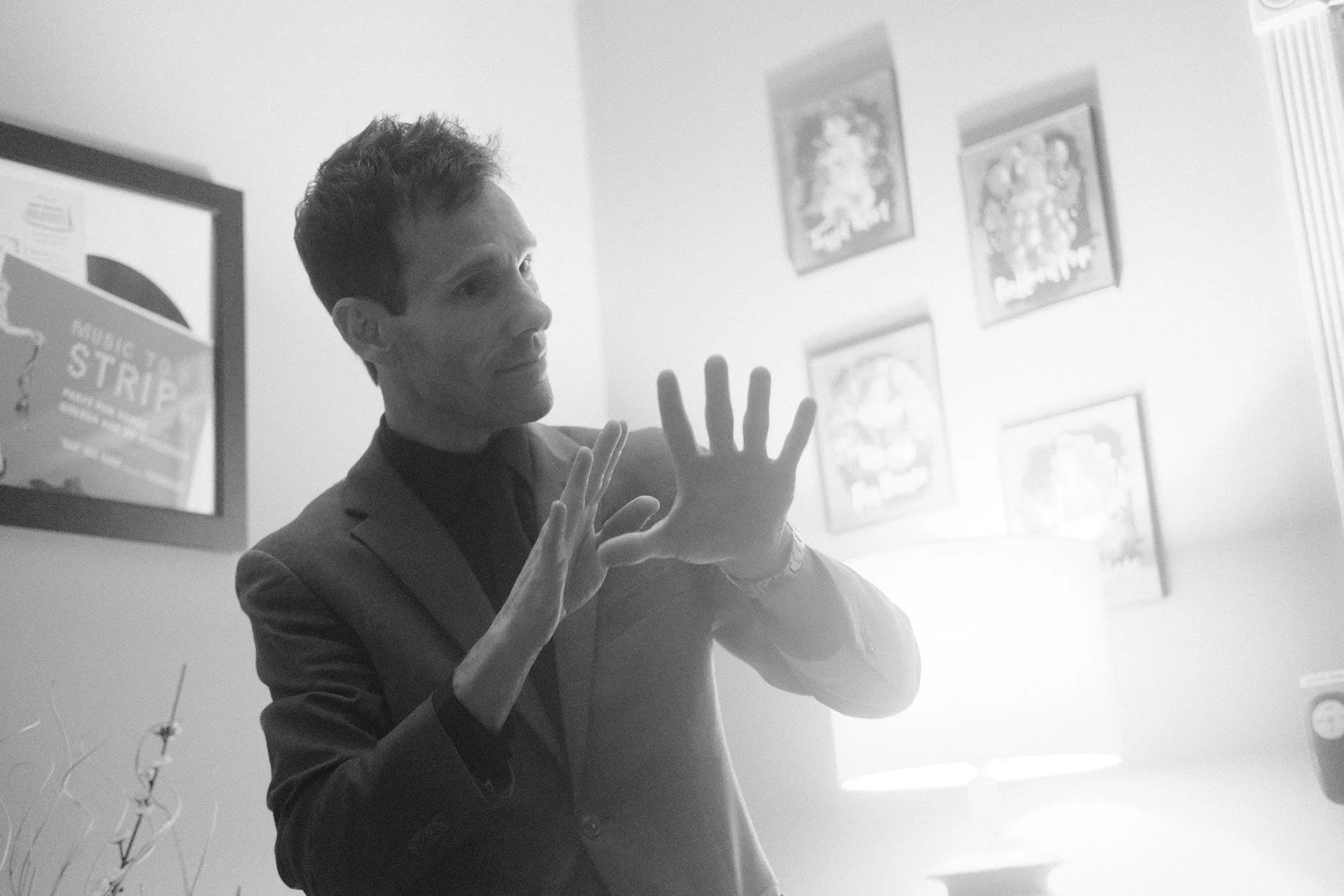 A man in a suit is looking at a mirror, with his hands raised and fingers spread, in a room decorated with framed pictures and posters on the wall.