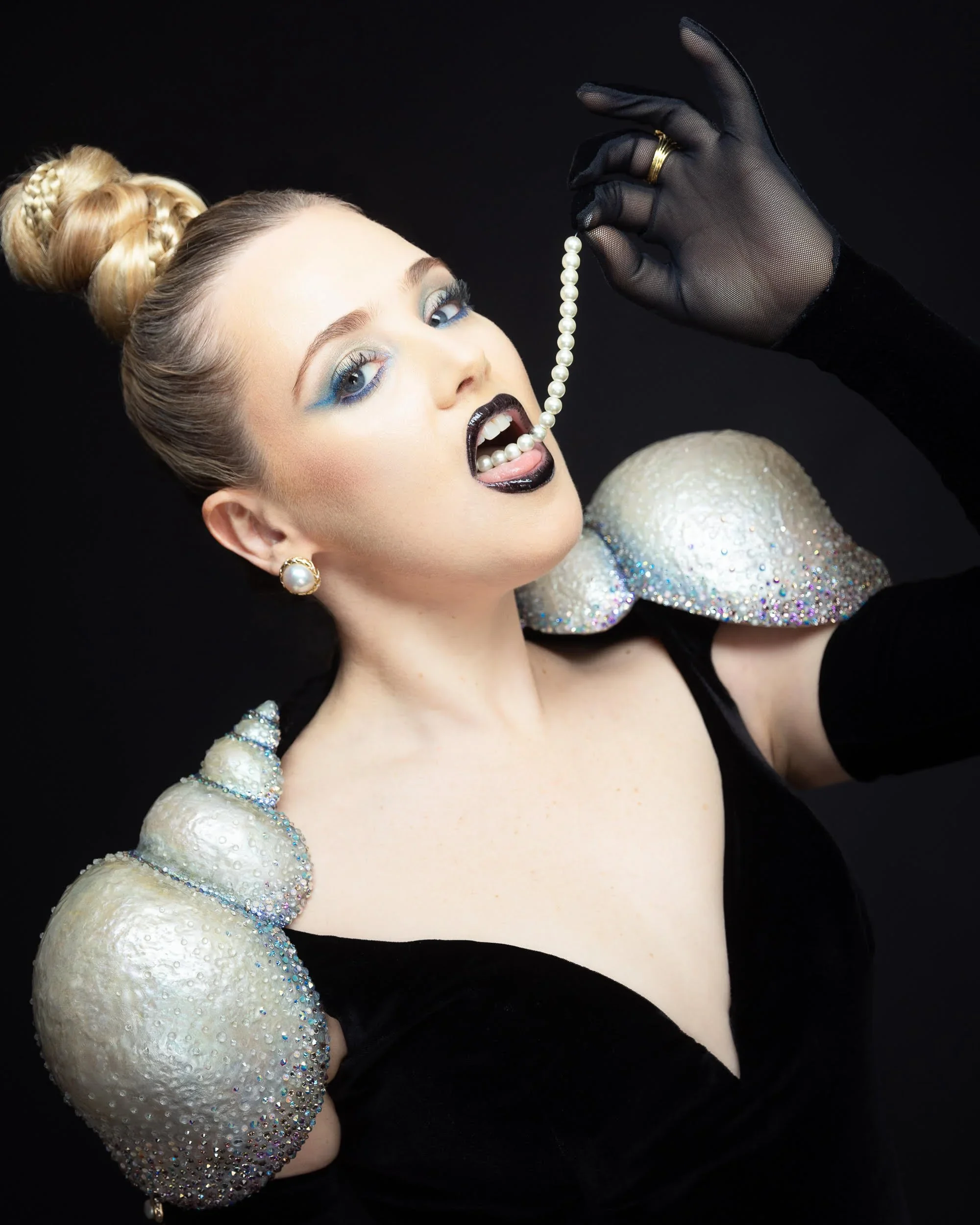A woman with blonde hair styled in an updo, wearing bold makeup with blue eyeshadow and black lipstick, dressed in a black outfit with sequin and pearl-embellished shoulder details, holding a strand of pearls inside her mouth, against a black background.