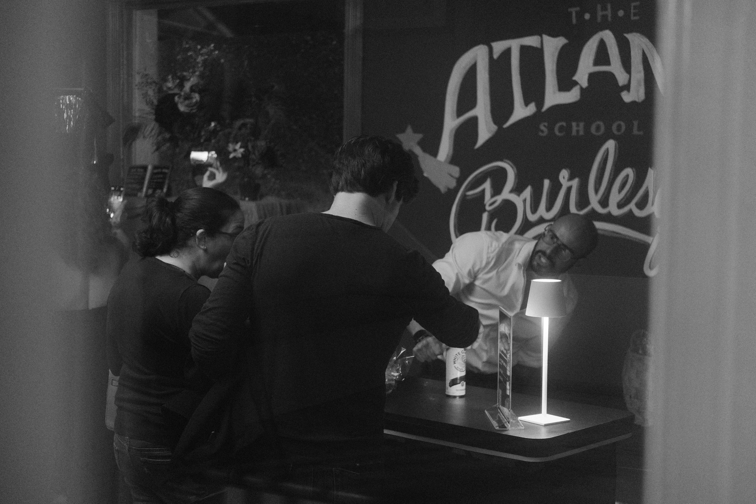 Three people are gathered around a table in a dimly lit room, with a lamp illuminating their work. Behind them, a large sign reads "The Atlantic School Burls". One man with glasses, wearing a white shirt, is leaning over the table, while a woman and