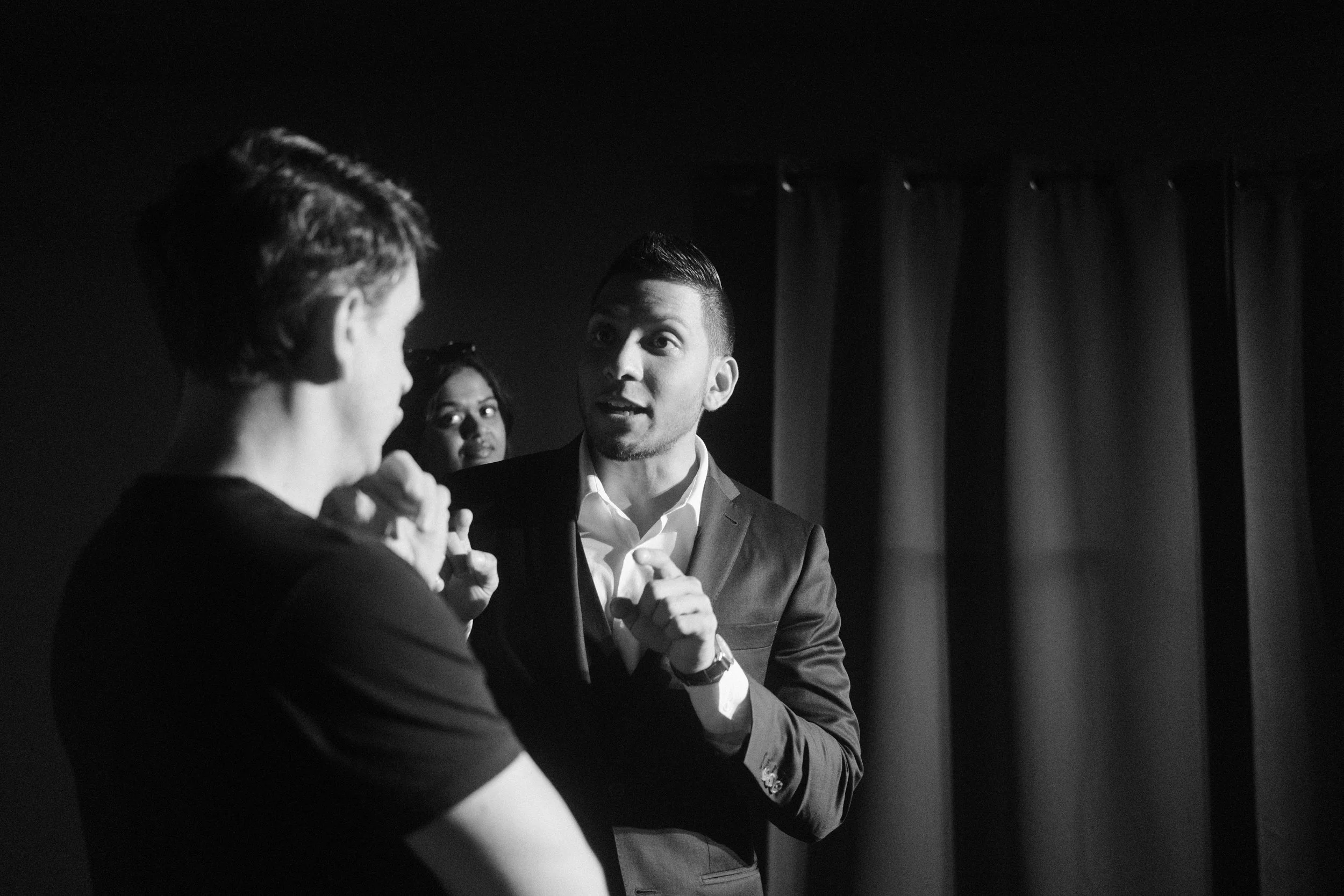 Black and white photo of a person in a suit talking to another person with short hair and a black shirt, with a woman observing in the background.