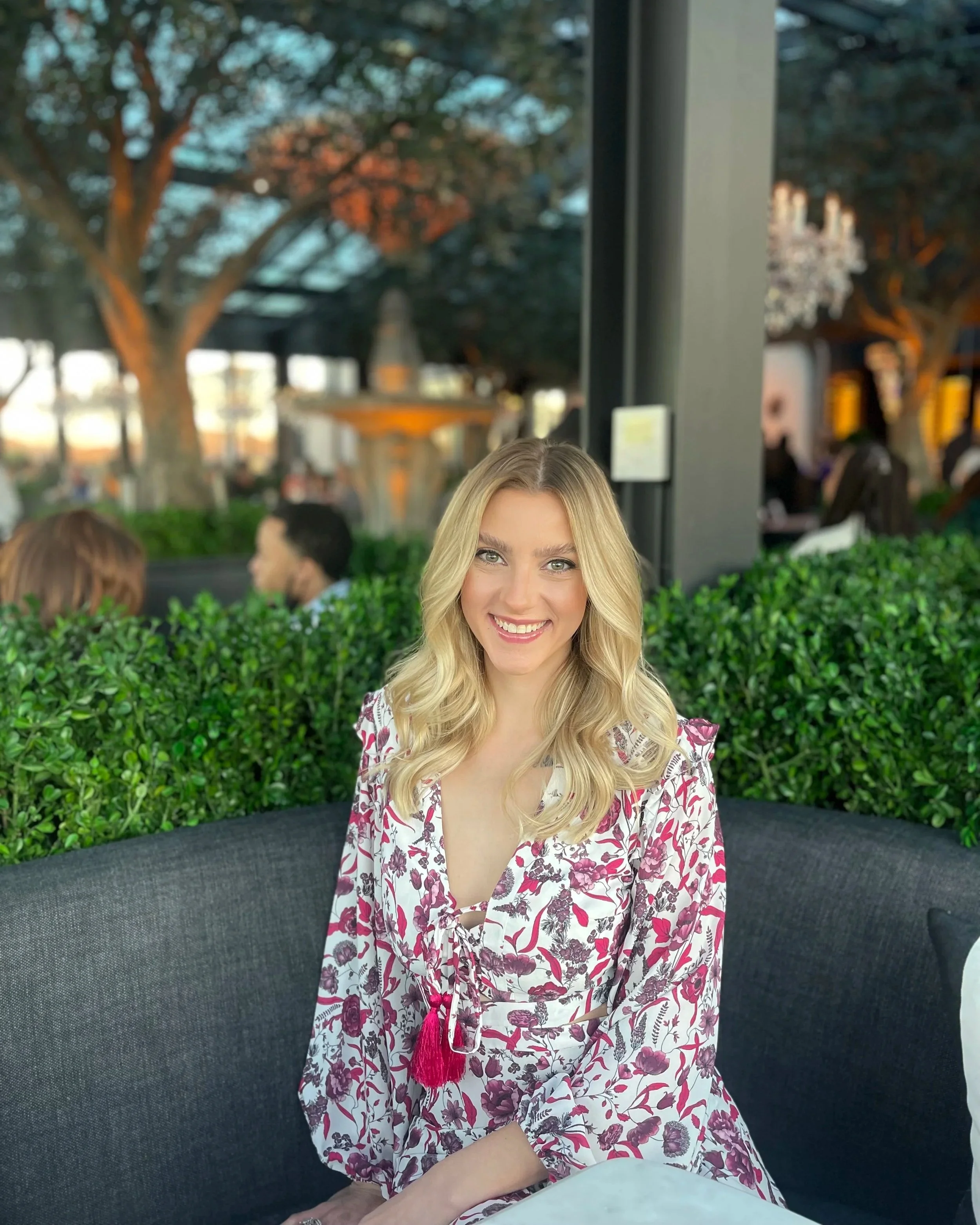A smiling woman with long blonde hair wearing a floral dress sitting at an outdoor cafe table with green bushes and trees in the background.