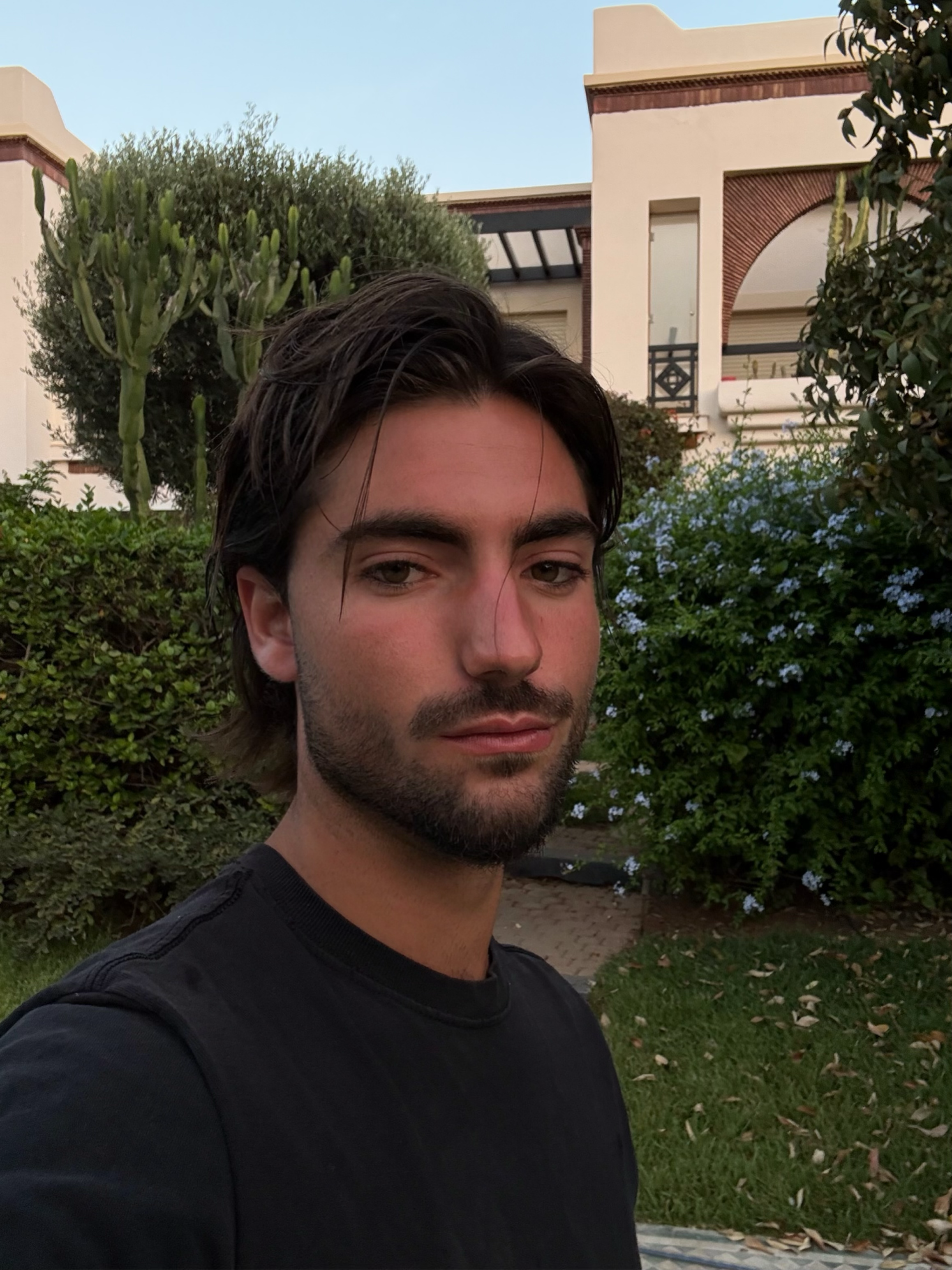 Un jeune homme aux cheveux bruns et à la barbe, portant un t-shirt noir, pose devant un bâtiment résidentiel avec des plantes et arbustes en arrière-plan.
