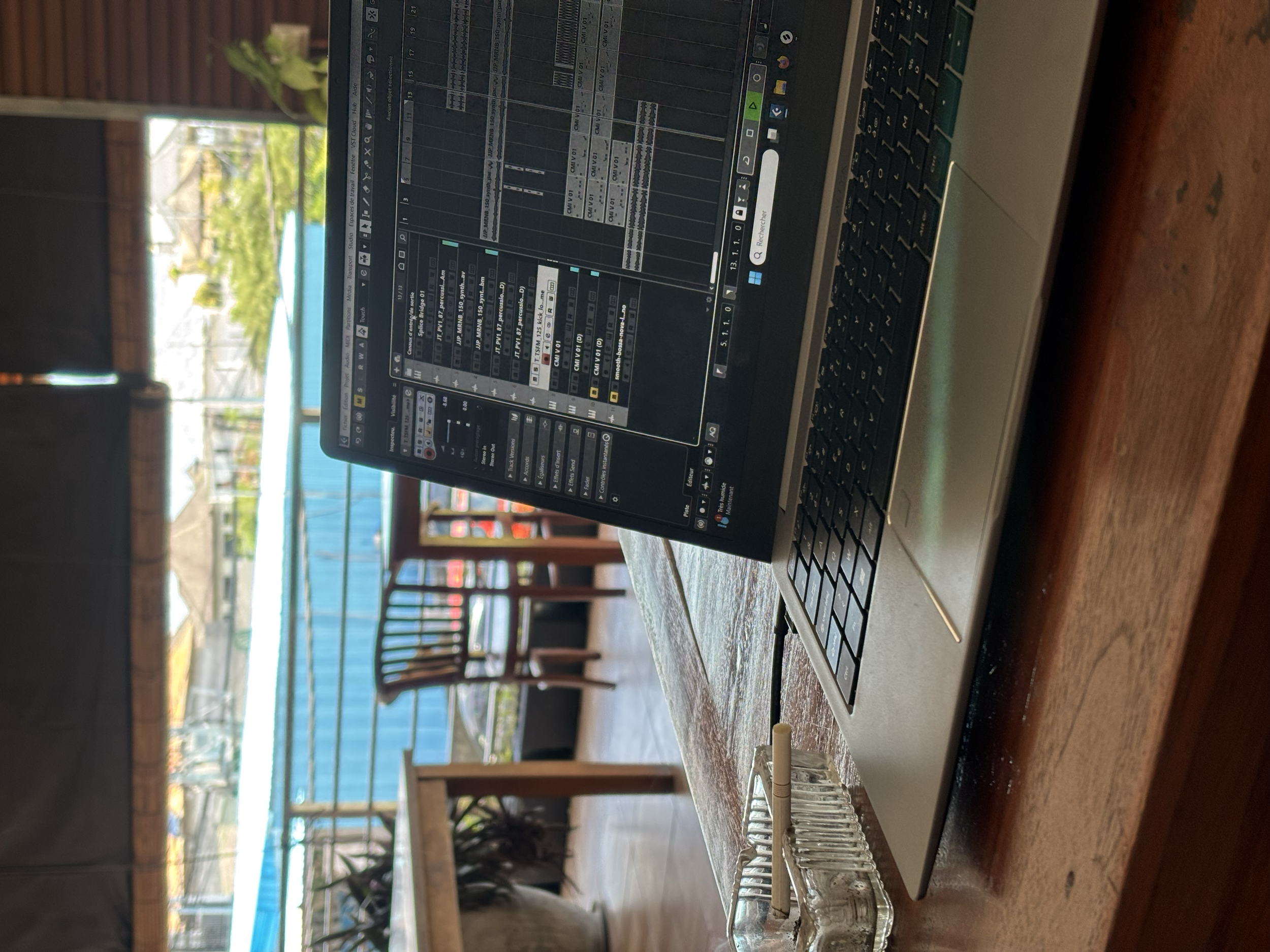 Un ordinateur portable affichant un logiciel de musique ou de DJ, placé sur une table en bois. En arrière-plan, une terrasse avec des chaises et une vue sur l'extérieur, avec une piscine et des plantes.
