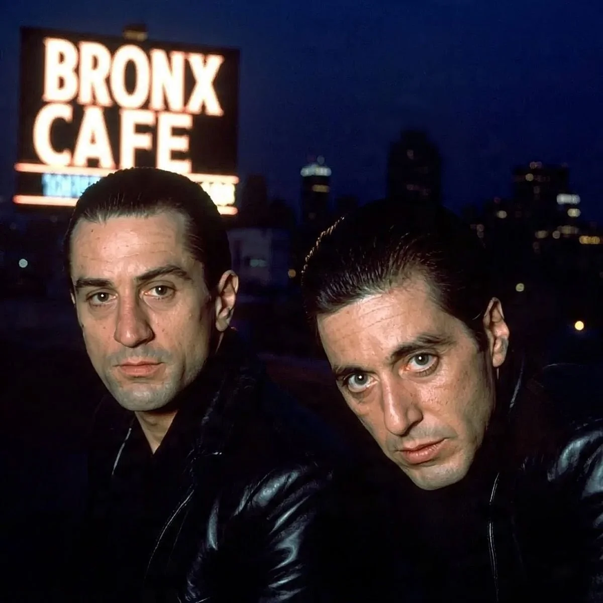 Deux hommes aux cheveux noirs et peau claire posent devant un fond nocturne avec un panneau lumineux indiquant 'Bronx Cafe' à New York.
